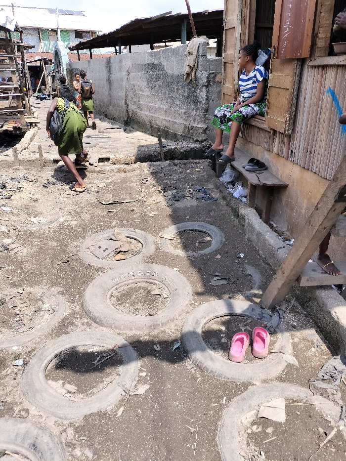 The scene depicts a narrow, unpaved alleyway within a densely populated area of Oworonshoki, Nigeria, under daylight conditions. The ground consists of uneven dirt, interspersed with numerous worn-out vehicle tires partially embedded, likely serving as stepping stones or ground stabilization. Various debris, including plastic bags and general refuse, is scattered along the path and in crevices.
On the right side, a building constructed from weathered wooden planks and other materials features an open window opening. A young woman is seated sideways within this opening, wearing a blue and white striped short-sleeved top and a patterned skirt. She has dark hair, possibly braided, and wears dark sandals on her feet. Her gaze is directed to the left side of the frame. Below her, a wooden bench holds a pair of black sandals and accumulated trash. A bare foot with an ankle chain is partially visible near the bottom right, next to a wooden support beam.
On the left side of the alley, a person, likely female, dressed in a green garment (possibly a wrapper) and carrying a black backpack, is walking away from the camera. Her feet appear to be wearing light-colored open-toed sandals or are bare. Further down the path, several other individuals, some also in green attire or carrying bags, are visible walking in the same direction.
In the mid-ground, separating the walking path from the right-side building, there is a low concrete block wall, exhibiting wear and discoloration. The background reveals additional residential or commercial structures with corrugated metal roofs. One roof in the far background appears to have solar panels installed. The overall environment suggests an informal settlement or community area characterized by rudimentary construction and visible refuse.