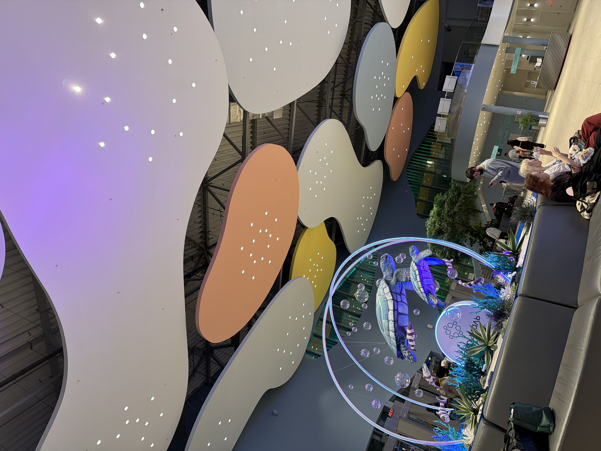 The image depicts the interior of what appears to be a public space, possibly a mall or airport, in Tyumen, Russia. The most striking feature is a decorative ceiling with large, irregularly shaped panels in colors such as white, orange, and yellow, each dotted with small lights. A decorative sculpture featuring turtles and bubble-like spheres, illuminated with blue and white light, hangs in the open space. The sculpture is accompanied by what looks to be artificial plants and signage in Russian.
Along the right side of the frame is a gray seating area where several people are sitting and interacting. A man in a gray shirt is standing, talking to a group of women seated on the couch. Other people are present in the background, further away, walking through the space. The lighting suggests a modern, public environment.