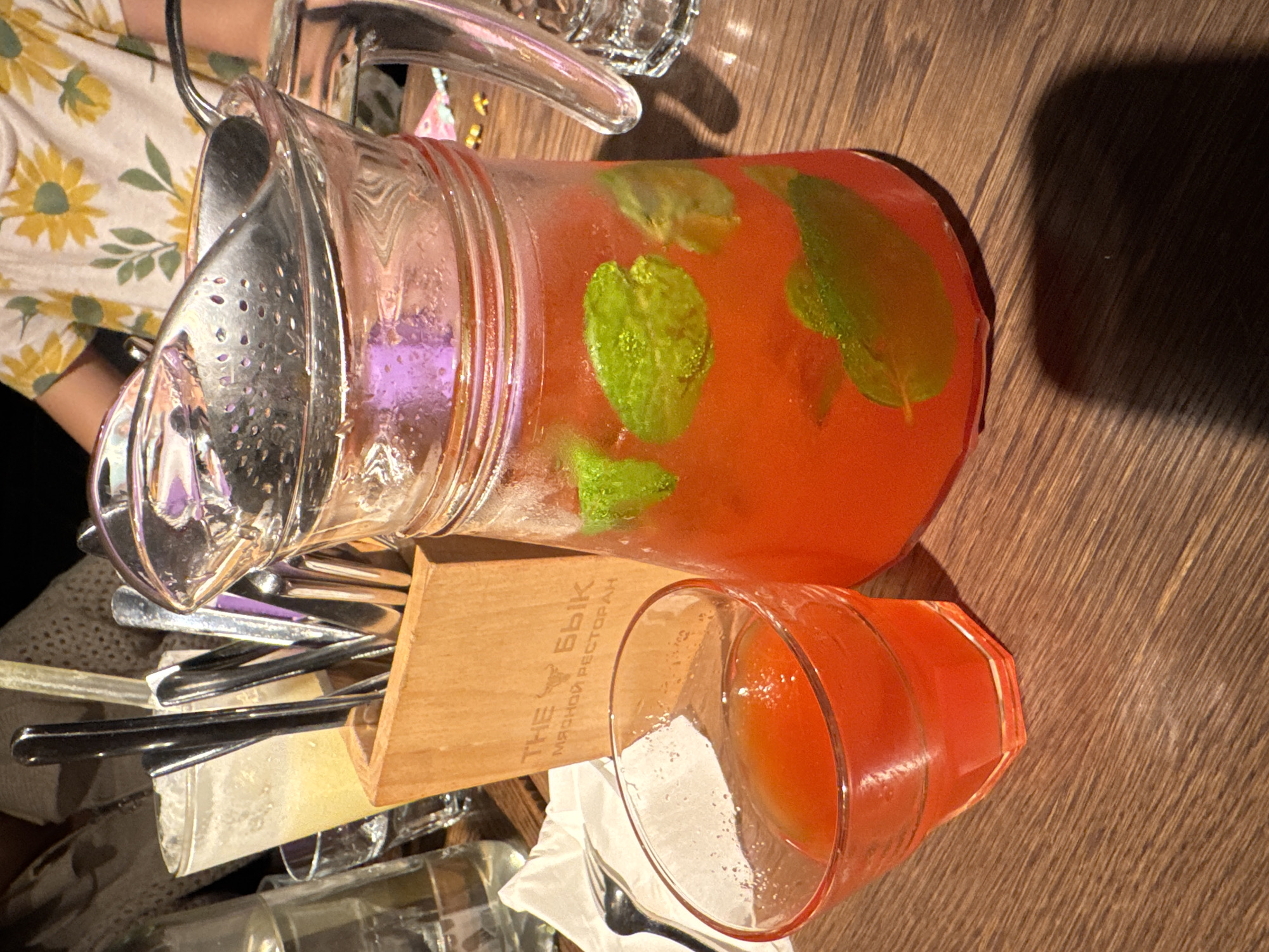 A high-angle, medium shot captures a wooden table at what appears to be "THE БЫК МЯСНОЙ РЕСТОРАН" (THE BULL MEAT RESTAURANT) in Saint Petersburg, Russia. In the foreground, a faceted glass contains a red beverage with ice and mint leaves. To its right, a large glass pitcher, also filled with a red beverage and mint, sits on the table. The pitcher has a metal strainer and a spoon resting inside. To the left of the pitcher, a wooden holder contains various cutlery, including spoons, forks, and knives. In the background, a person wearing a yellow t-shirt with a floral pattern is partially visible. The lighting suggests an indoor setting with artificial light.