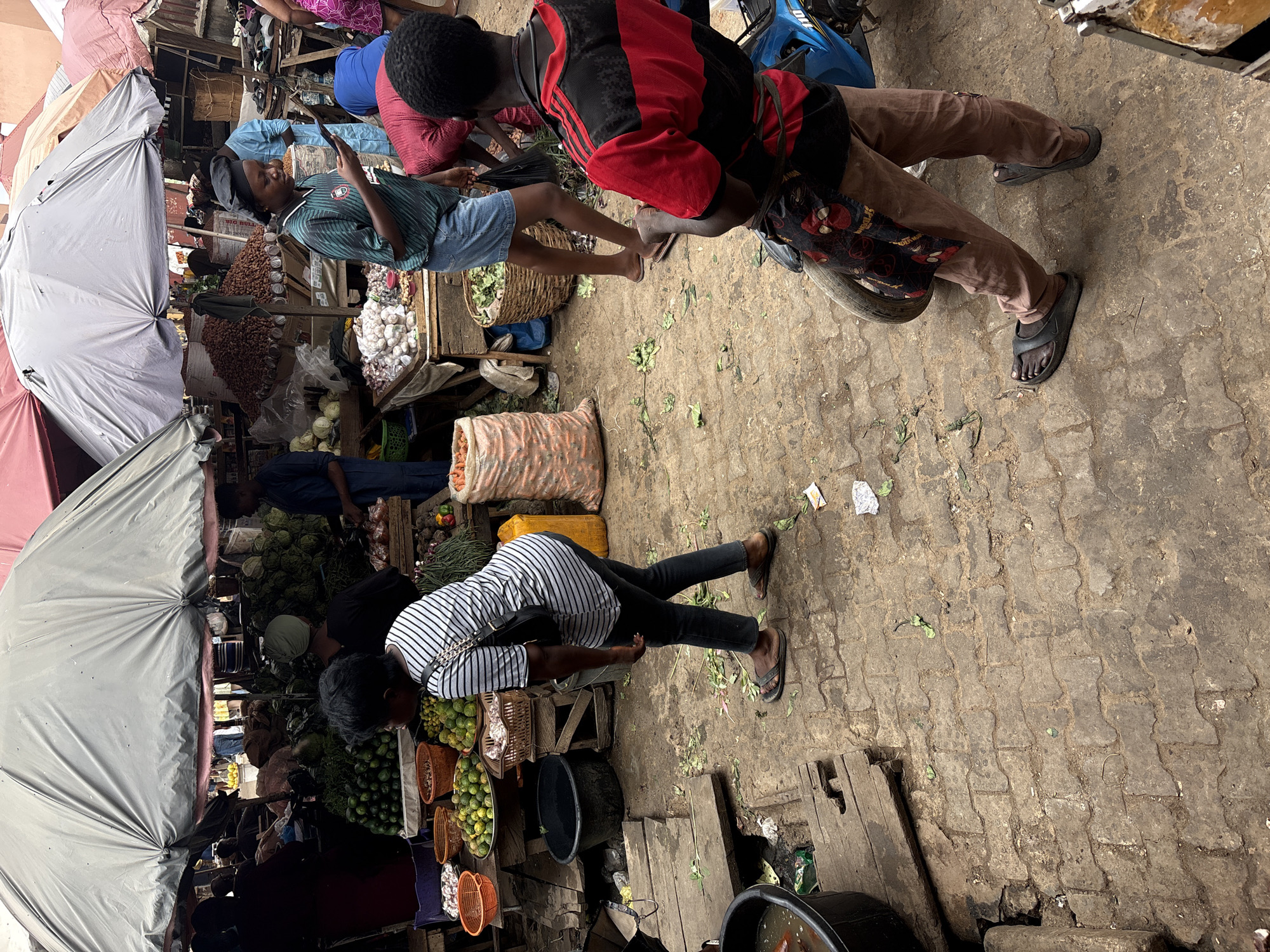 The image depicts a bustling marketplace in Lugbe, Nigeria. The scene is dominated by vendors and shoppers navigating a crowded, open-air space. A white umbrella shields a stall laden with produce, including leafy greens, limes, and other vegetables. Shoppers are interacting with vendors; some are examining goods while others appear to be in the process of purchasing items. One person is riding a scooter. The ground is a mix of paved areas with scattered debris, including leaves and bits of trash.
