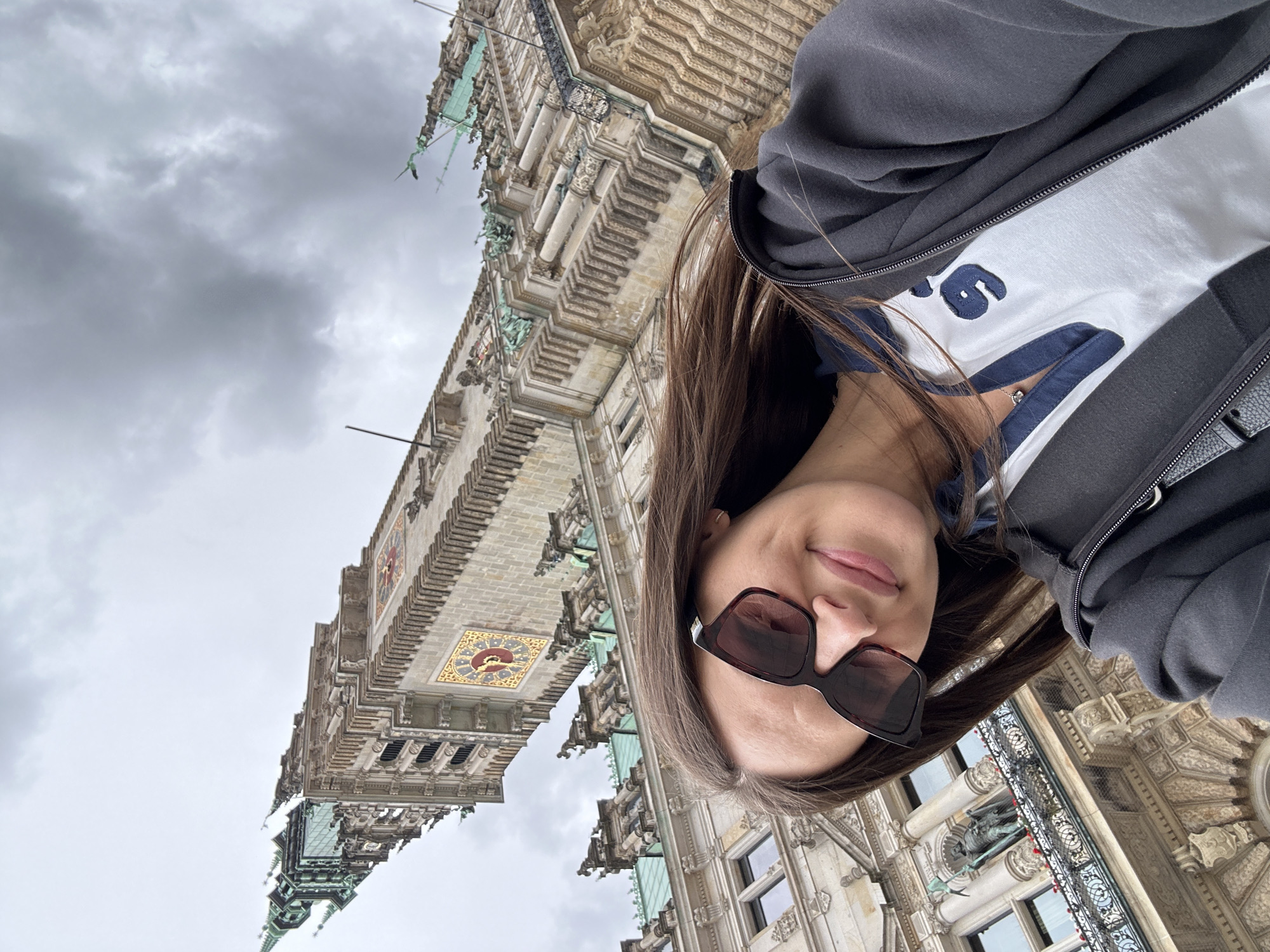 The image is a selfie, rotated 90 degrees counter-clockwise, featuring a young woman against the backdrop of a grand, ornate building under an overcast sky.
**Subjects:**
The main subject is a young woman with long, straight dark brown hair, seen from the chest up. She is wearing dark, square-framed sunglasses, a white t-shirt, and a dark gray zip-up hoodie. A delicate silver-toned necklace with a small, round pendant is visible around her neck.
The background is dominated by a majestic, multi-story stone building, exhibiting detailed Neo-Renaissance architecture. It features intricate carvings, numerous windows, balconies with ornate railings, and a prominent clock tower. The clock face is square with gilded embellishments. Parts of the building's roof or spires show green patination, suggesting copper or bronze elements.
**Activity or event taking place:**
The image captures a selfie, with the woman posing in front of the impressive architectural landmark.
**Notable details:**
The sky is entirely overcast with thick gray clouds, indicating a cloudy or dull day. The building's distinctive architecture, particularly the clock tower and overall design, strongly resembles the Hamburg Rathaus (City Hall) in Hamburg, Germany. The woman has a calm, slightly smiling expression.
**Visible text:**
A dark blue, possibly embroidered or printed, design is partially visible on the white t-shirt, resembling the numeral "6".