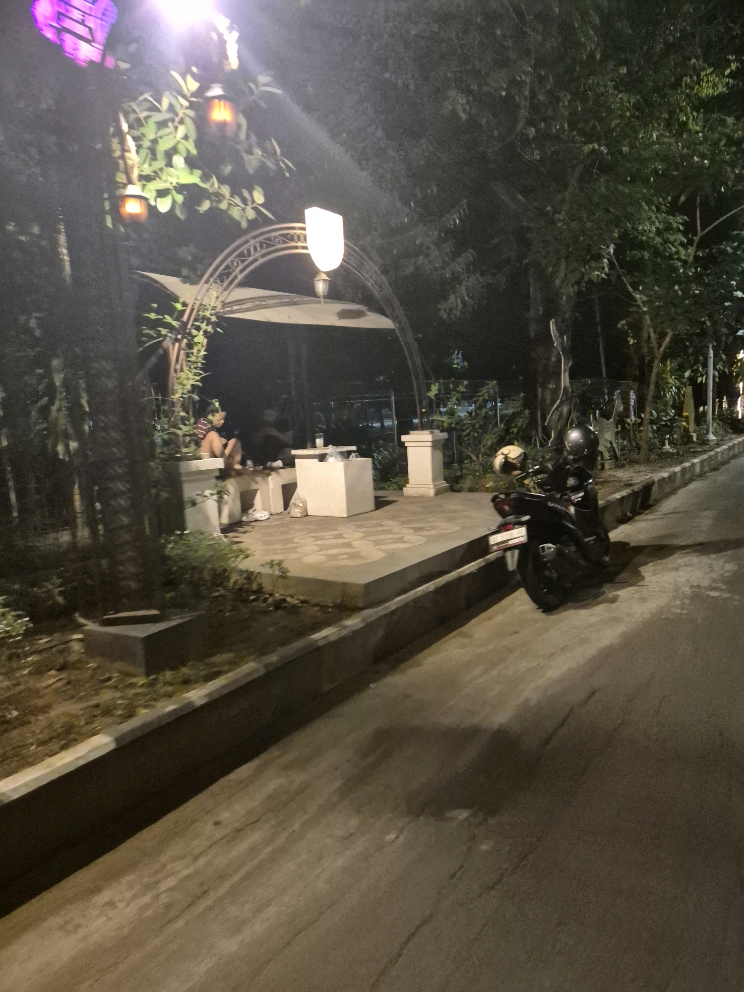 The image depicts an outdoor seating area at night, possibly a cafe or restaurant, located in Surakarta, Indonesia. In the foreground, a dark motorcycle is parked on the right side of a concrete pathway. The motorcycle has two helmets on its seat. To the left of the motorcycle, a concrete curb separates the pathway from a grassy area.
The background reveals a covered seating area with arched metalwork. Inside the covered area, two individuals are seated on a concrete bench. One person, wearing a striped shirt and dark shorts, is facing away from the camera. Another person is partially visible behind them. A white table sits in front of them with drinks and a plastic bag.
Decorative lanterns, one emitting a warm orange glow and another a brighter white light, are visible hanging from trees and the structure, illuminating the scene. A large, purple, illuminated decorative object is also present in the upper left corner of the image. The overall environment suggests a relaxed, evening atmosphere with ambient lighting.