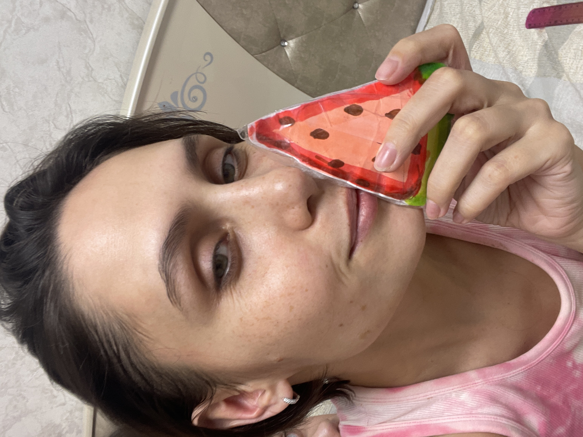 A woman is holding a watermelon-shaped object up to her face. Her hair is dark and pulled back. She has freckles on her face and is wearing a small earring. She is wearing a pink and white tank top. The background includes a headboard with buttons and a white wall. The location is not mentioned in the image, and it cannot be reliably determined from the image alone.