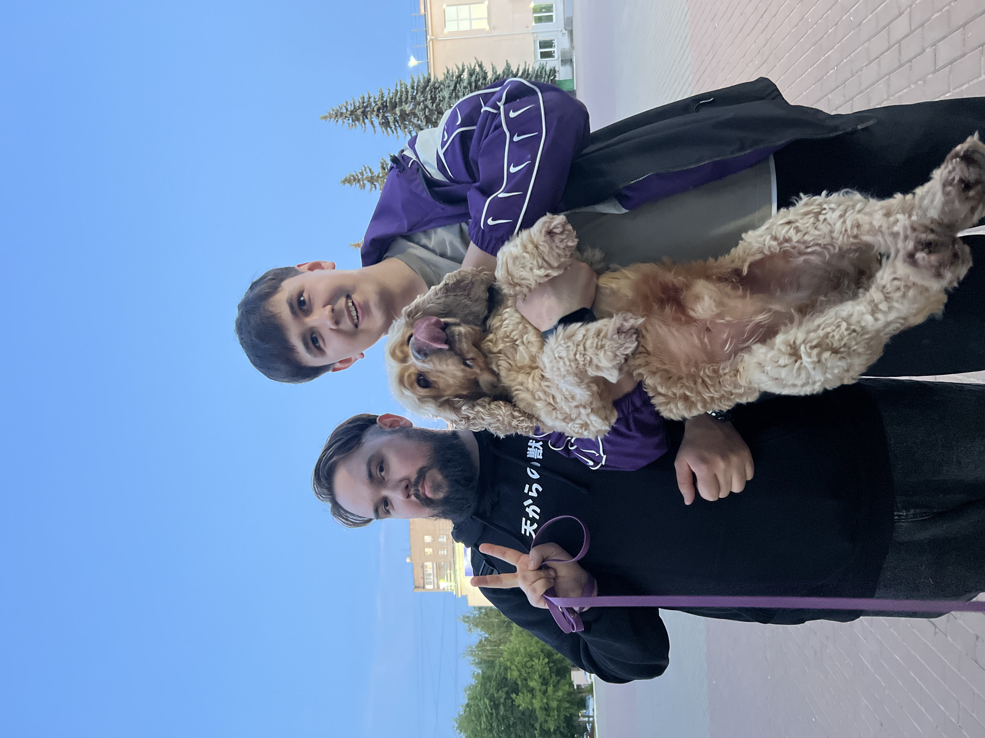 This outdoor image captures two men and a dog posing under a clear blue sky in Chaikovsky, Russia. The photo appears to be taken from a slightly low angle.
On the left, a younger man with short dark hair smiles broadly while holding a fluffy, light brown dog upside down. The dog, possibly a Cocker Spaniel, has its tongue out and appears playful. The young man wears a grey t-shirt beneath a purple and white track jacket, which is adorned with multiple white Nike swoosh logos down the sleeves.
To his right, an older man with a dark beard and dark hair looks towards the camera. He wears a black hoodie with white Japanese text on the chest, which reads "天からの贈り物" (Ten kara no okurimono), translating to "gift from heaven." He makes a peace sign with his right hand and holds a purple leash, which extends downwards.
The background features several evergreen trees and light-colored buildings, some with visible windows and brickwork, suggesting an urban or suburban setting. The bright, clear blue sky indicates fair weather and daytime. The overall mood of the scene is cheerful and friendly, with both people and the dog looking happy and relaxed.