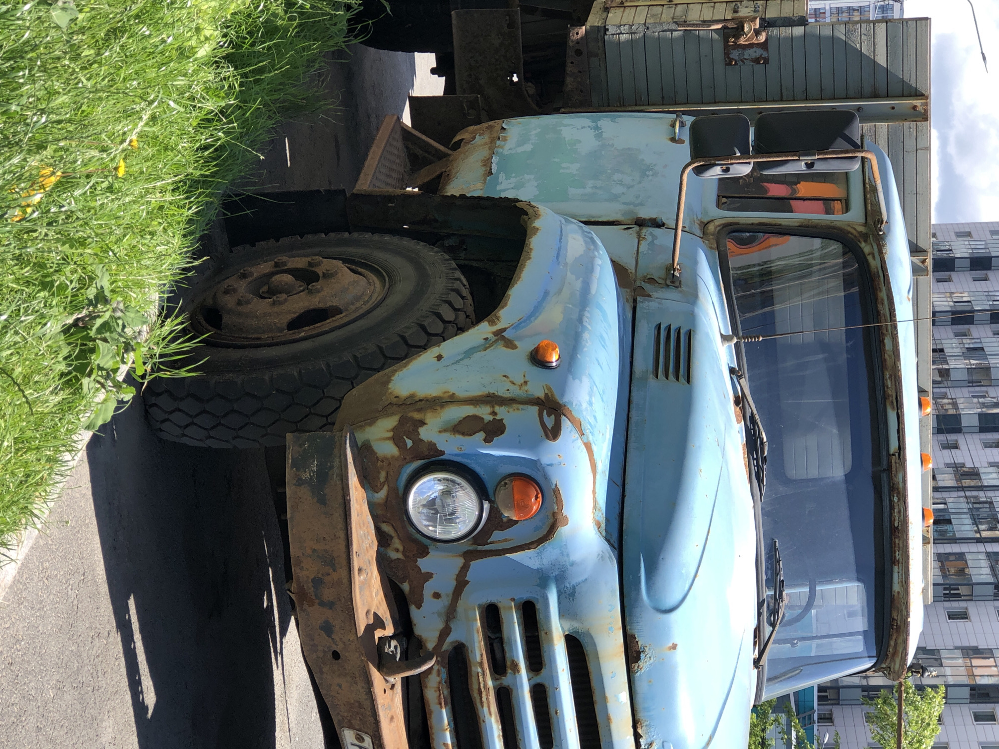 The image shows a close-up, slightly angled view of a heavily rusted, old blue truck parked on an asphalt surface next to a patch of green grass. The truck is a Soviet-era model, likely from the 1970s or 1980s, with a weathered wooden cargo bed. The paint is peeling, revealing significant rust on the hood, fenders, and grille. A round headlight with an orange turn signal above it is visible on the front right fender. The truck has large, rugged tires. In the background, a modern, multi-story apartment building with numerous windows can be seen, suggesting an urban setting. The sky is partly cloudy. The overall impression is one of decay and abandonment amidst modern urban development. The scene is consistent with the industrial or residential outskirts of Saint Petersburg, Russia, where older infrastructure often coexists with newer construction. There are no people or animals visible in the image. The lighting suggests it is daytime, likely midday, with sunlight casting shadows on the ground.