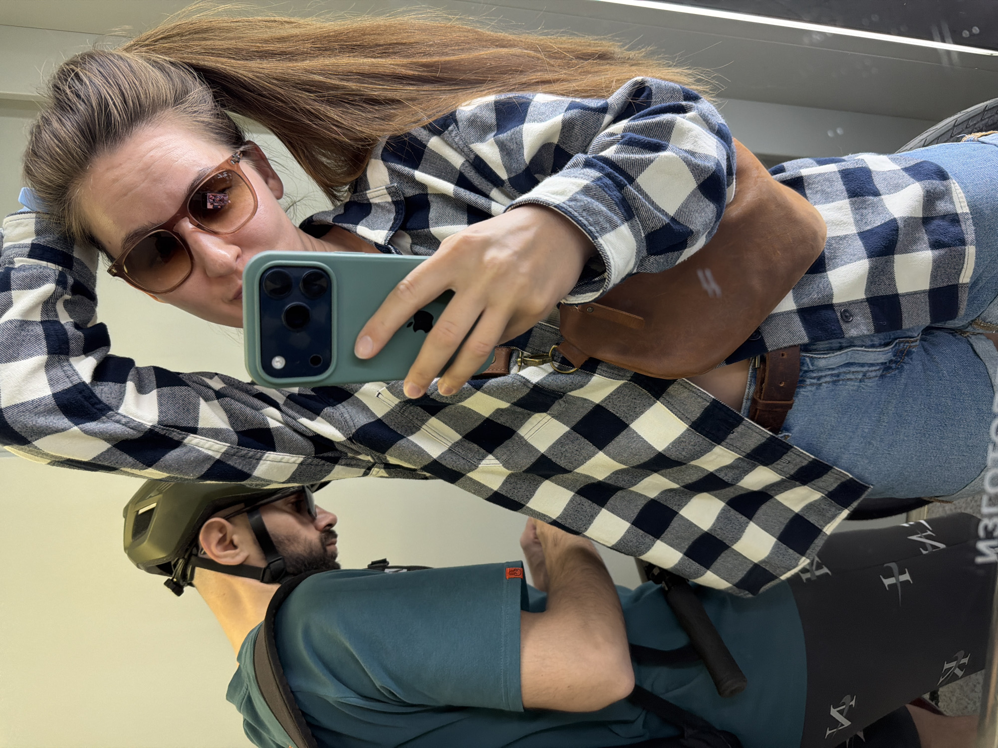 A woman with long brown hair pulled back into a high ponytail and wearing sunglasses, a blue and white plaid shirt, a tan leather fanny pack, and blue jeans, is taking a selfie with a green smartphone in a mirrored surface. The reflection shows a man wearing a green helmet and a blue-green t-shirt. The man appears to be holding a bicycle handle. The location is in Tyumen, Russia, as the text on the reflection of the bicycle gear is written in Russian.