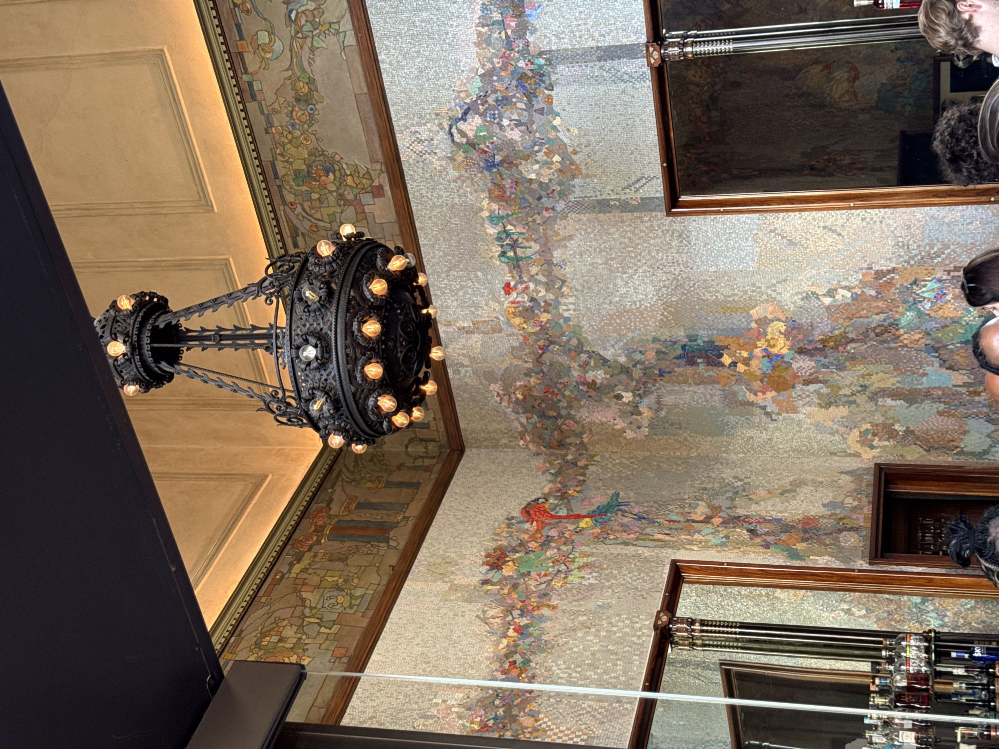 This image captures the interior of the Camparino in Galleria, located in Milan, Italy. The scene features the bar's distinctive Art Nouveau aesthetic, dominated by intricate mosaic artwork and an ornate chandelier.
The walls are largely covered in a mosaic of small, light-colored tiles, over which vibrant, fragmented mosaics depict stylized flora and fauna, including red, green, and blue parrots, appearing to "drip" or flow down from a central horizontal band. Above this, a richly colored mosaic frieze encircles the room, displaying more traditional motifs of foliage, abstract shapes, and architectural elements in muted greens, browns, reds, and blues.
The ceiling features a recessed section above the frieze, illuminated by warm, hidden lighting. Above this, the ceiling itself is a warm beige or light gold color with decorative rectangular molding.
A large, dark, wrought-iron chandelier hangs prominently from the center of the ceiling. It is multi-tiered and adorned with numerous exposed, glowing incandescent light bulbs that cast a warm, yellowish light throughout the space.
In the lower left, a section of a bar is visible, showcasing multiple bottles of liquor on shelves, reflected in a tall, dark-framed mirror. Another dark-framed mirror is partially visible on the right. The top of several individuals' heads is visible at the bottom of the frame, indicating the presence of patrons in the establishment.
