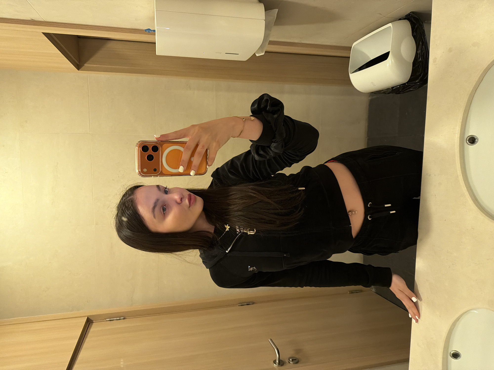 A young woman is depicted taking a mirror selfie in a public restroom, likely located in Sofia, Bulgaria. She has long, dark hair and light eyes, and wears a black velour tracksuit, comprising a zippered hoodie and matching sweatpants. A gold-colored logo, possibly Juicy Couture, is visible on the left side of her hoodie. She also wears a delicate gold bracelet on her left wrist and has a small, beaded navel piercing. Her fingernails are painted white.
She is holding an orange-cased smartphone, with visible camera lenses and a white MagSafe ring, to capture her reflection. The setting features light beige tiled walls, light wooden panels or doors, and a light-colored countertop with a partial view of a white sink. Above her head, a white paper towel dispenser is mounted on the wall. To her right, a white waste bin, lined with a black plastic bag, is visible on the countertop. The lighting is artificial and bright, suggesting an indoor environment. The woman maintains a neutral expression, looking directly into the phone's camera.