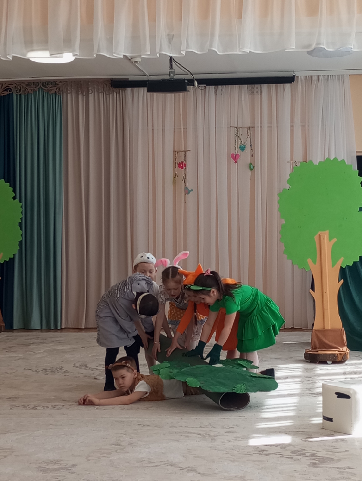 This image captures an indoor children's performance, likely a play or skit, featuring six young children dressed in animal-themed costumes. The scene is set on a light-colored, patterned carpeted floor, against a backdrop of light sheer curtains layered over heavier teal and beige curtains, suggestive of a stage or performance area in a school or kindergarten in Izhevsk, Russia.
In the foreground, a child in a brown, fur-like costume with small ears is lying prone on the floor, looking to the right with a focused expression. Grouped around a green, log-shaped prop are four other children. One child, wearing a grey patterned costume and a white cap, is leaning over the prop. Adjacent to them, a child with pink bunny ears and an orange patterned dress, along with another child sporting orange fox ears and an orange outfit, are also bent over the green prop. A fifth child in a bright green dress and dark gloves is actively engaging with the prop. A sixth child with bunny ears is partially visible behind the child in the grey costume.
The stage is minimally decorated with two large, flat cardboard trees with green foliage and brown trunks, positioned on either side of the stage. Small decorative hearts and flowers are suspended from wires in the background. Overhead, a rectangular light fixture and a black speaker or projector are visible. The children appear engrossed in their roles, participating in what seems to be a playful and imaginative theatrical event. No visible text is present in the image.