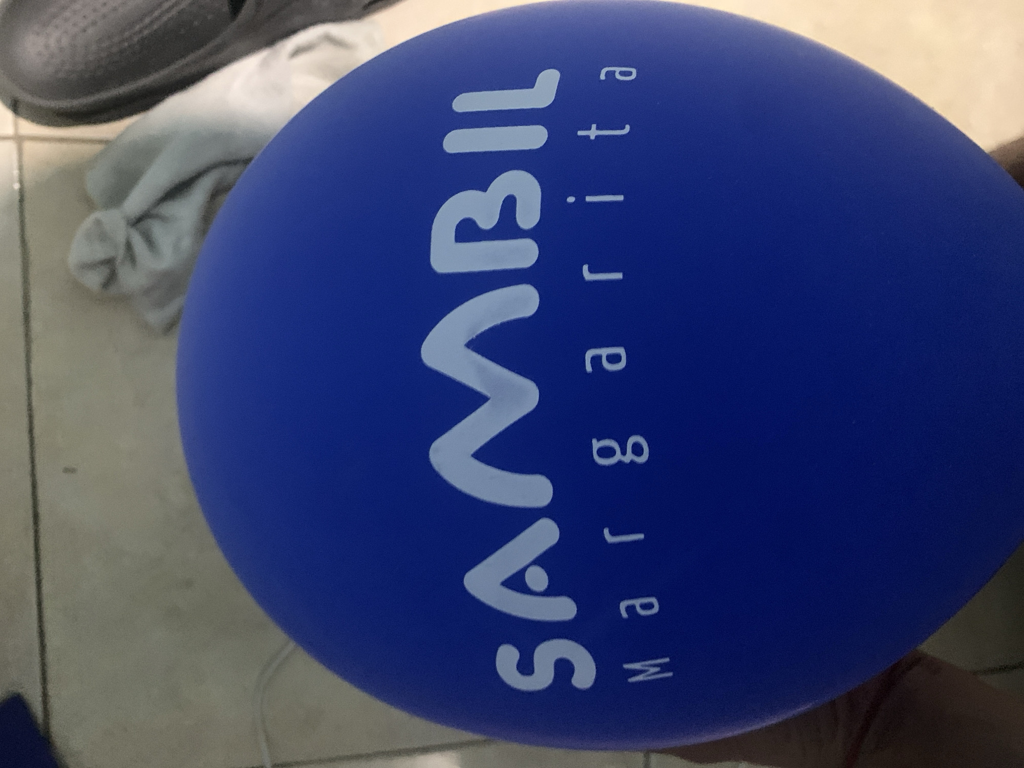 A close-up image captures a large, inflated blue balloon positioned prominently in the foreground, partially obscuring other items. White text is emblazoned on the balloon, reading "SAMBIL" horizontally in a stylized font, with "Margarita" written vertically below it, aligned to the right of "SAMBIL". The balloon itself takes up a significant portion of the frame, tilted on its side.
In the upper left quadrant of the image, the toe and part of the upper surface of a dark grey, possibly rubber, shoe or sandal are visible. Beneath the shoe and partially beneath the balloon, a crumpled light-colored garment or cloth, likely white or off-white, rests on a tiled floor. The floor appears to be made of light-toned, possibly grey or beige, square tiles, with visible grout lines. The lighting suggests an indoor setting. No specific activity or event is taking place; the items are stationary. The visible text "SAMBIL Margarita" indicates a connection to Sambil shopping malls, specifically the one located on Margarita Island, which aligns with the provided location context of Santa Ana, Venezuela, a city on Margarita Island.