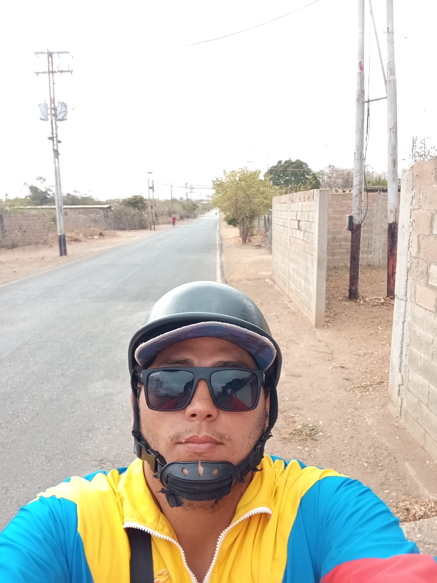 A selfie of a person wearing a helmet and sunglasses on a road with an asphalt surface. The person is also wearing a jacket with blue and yellow colors. The road is flanked by walls and utility poles. The location is Juan Griego, Venezuela.