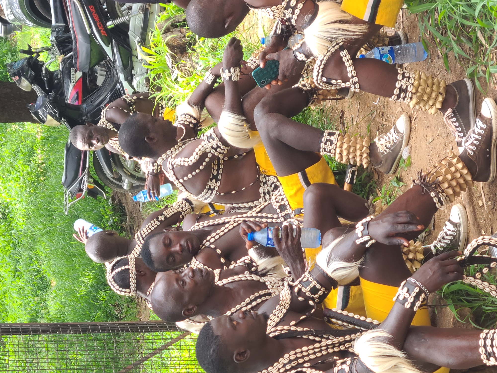The image captures a group of men adorned in traditional attire, including cowrie shell necklaces and bracelets, and what appear to be raffia-like leg adornments. They are seated outdoors on what looks like a dirt path surrounded by green grass and foliage, with a large tree trunk visible in the background. Two motorcycles are parked behind the men, one of which has "JINCHENG" branding. The men are dressed in yellow shorts and some are wearing sneakers. Some individuals are holding bottles of water, and one man is looking at a mobile phone. The scene suggests a cultural gathering or performance. The specific location within Jos, Nigeria, cannot be determined from the visual information alone.