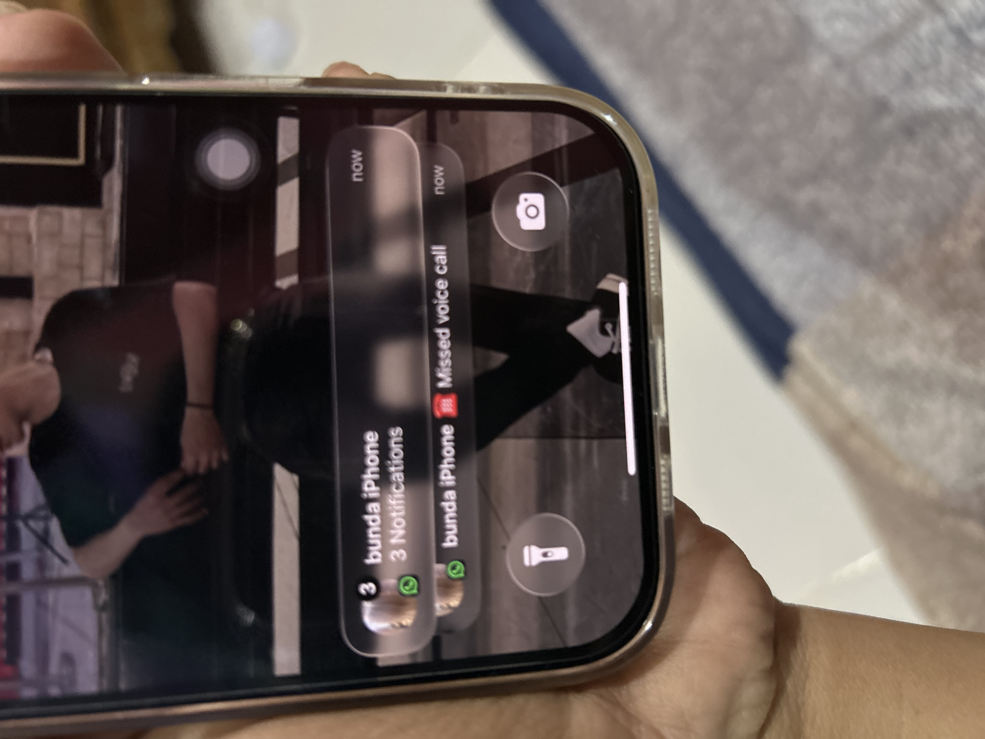 In the city of Depok, Indonesia, a person's hand holds a smartphone, displaying a lock screen with notifications. The dominant notification is from "bunda iPhone" indicating "3 Notifications" and another "Missed voice call" also from "bunda iPhone." Below these notifications, two circular icons are visible: a flashlight icon on the left and a camera icon on the right. The background of the phone screen shows a partially visible image of a person wearing a black t-shirt, with their arms resting on a surface. The overall scene appears to be an indoor setting, as suggested by the lighting and the background elements.