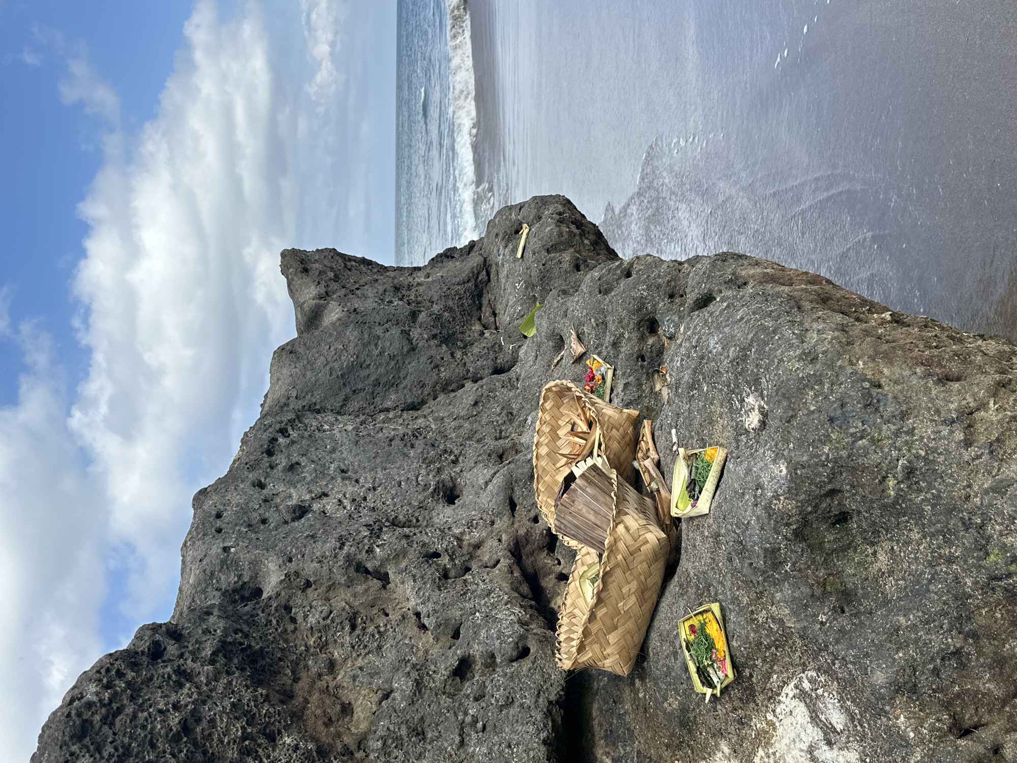 On a rocky outcropping on a beach near North Kuta, Indonesia, a woven basket and two small trays are visible. The trays contain flower petals and greenery. A dark sand beach is in the background, and the sky is partly cloudy.