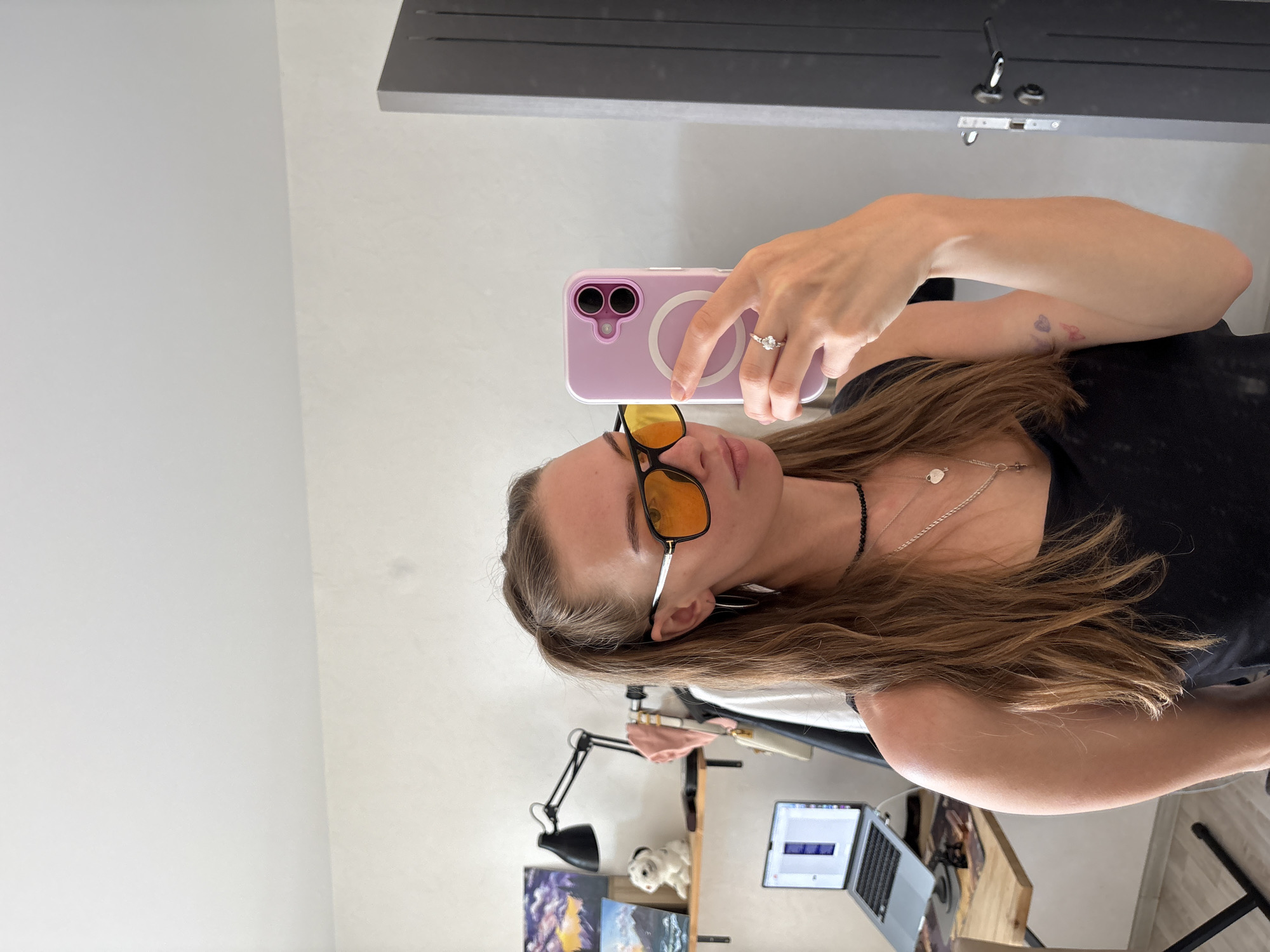 A woman is taking a mirror selfie in an indoor environment. She has long, brown hair pulled into a ponytail and is wearing a black tank top, sunglasses with orange lenses, a beaded choker, and a silver necklace. A ring is visible on her right hand, which is holding a light purple phone. In the background, a shelf holds two framed pictures, a desk lamp, and a white figurine of a dog. A laptop is on a desk to the left of the woman. A dark gray door is visible to the right.