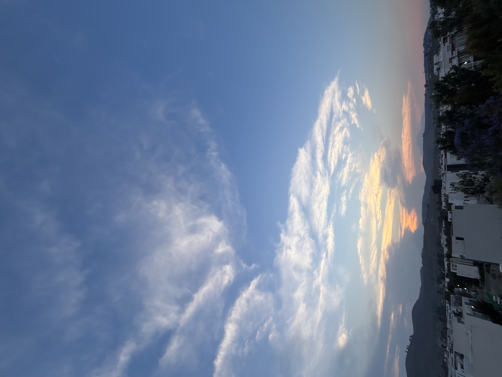The image captures a sky with a gradient of blues and whites, transitioning towards orange and yellow hues near the horizon. The sky is filled with scattered clouds, varying in shape and density. The horizon shows the presence of a mountain range. In the foreground, there are buildings and trees, suggesting an urban or suburban environment. The exact location is in Santa Clara Ocoyucan, Mexico. The image appears to be a landscape photograph with a focus on the sky and the natural elements, with some indication of human habitation.