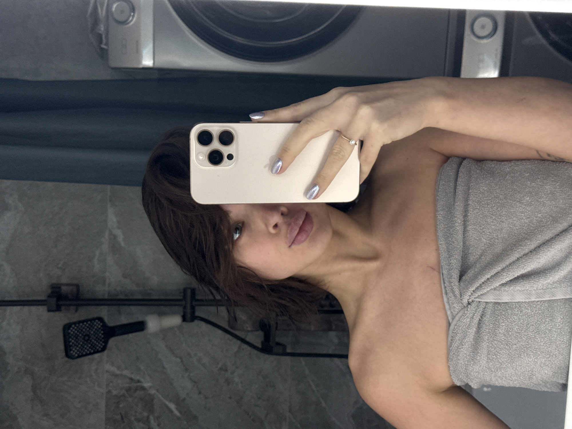 A woman is taking a selfie in a bathroom. She is holding a smartphone in front of her face, partially obscuring it. She has shoulder-length brown hair and is wrapped in a gray towel. The bathroom has a gray marble wall with a black shower head and fixture. A washing machine and dryer are visible in the upper portion of the image, reflected in a mirror. There is no information to suggest this image was taken in the city of Лобаново, Russia.