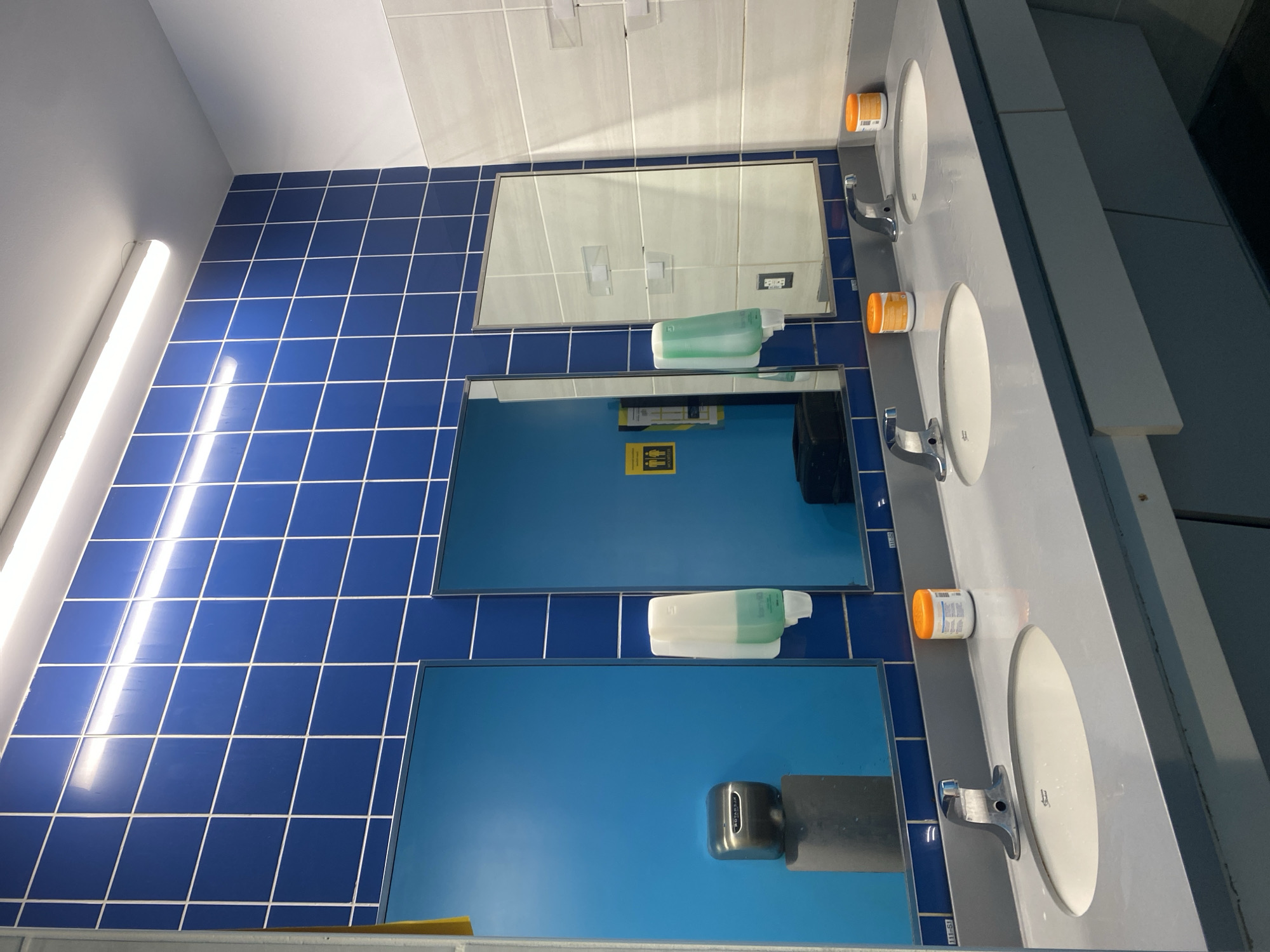 The image shows a public restroom in Virginia Beach, United States of America. The restroom features a row of three sinks with grey countertops and white porcelain basins. Each sink has a chrome faucet. On the countertops, there are two orange-lidded containers of what appears to be lotion or sanitizer. Behind the sinks, a wall is tiled with blue square tiles and white grout.
To the left, there is a bright blue panel with a silver hand dryer mounted on a grey cylindrical pedestal. Above the blue panel, a yellow and black sign with pictograms of a man and a woman is visible. Adjacent to this, a white soap dispenser is mounted on the blue panel.
In the center, a mirror reflects a blue door and a dark object on the floor. Another white soap dispenser is mounted on the blue panel to the right of the mirror. To the far right, a white tiled wall section is visible, with some clear plastic items mounted on it. The overall lighting is provided by fluorescent lights running along the ceiling.