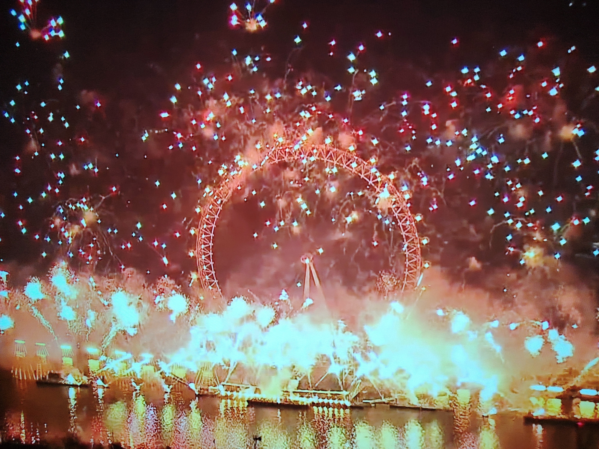 The image captures a spectacular fireworks display over a body of water, with the London Eye ferris wheel prominently featured in the background. Numerous bursts of colorful fireworks, primarily red and blue, fill the night sky, creating a festive and dynamic atmosphere. The London Eye, illuminated and outlined in red, stands as a central landmark amidst the pyrotechnics. In the foreground, structures along the waterfront, likely part of a celebratory event, are aglow with bright lights, casting reflections on the water's surface. The overall scene suggests a significant public event or celebration taking place in London, United Kingdom.
