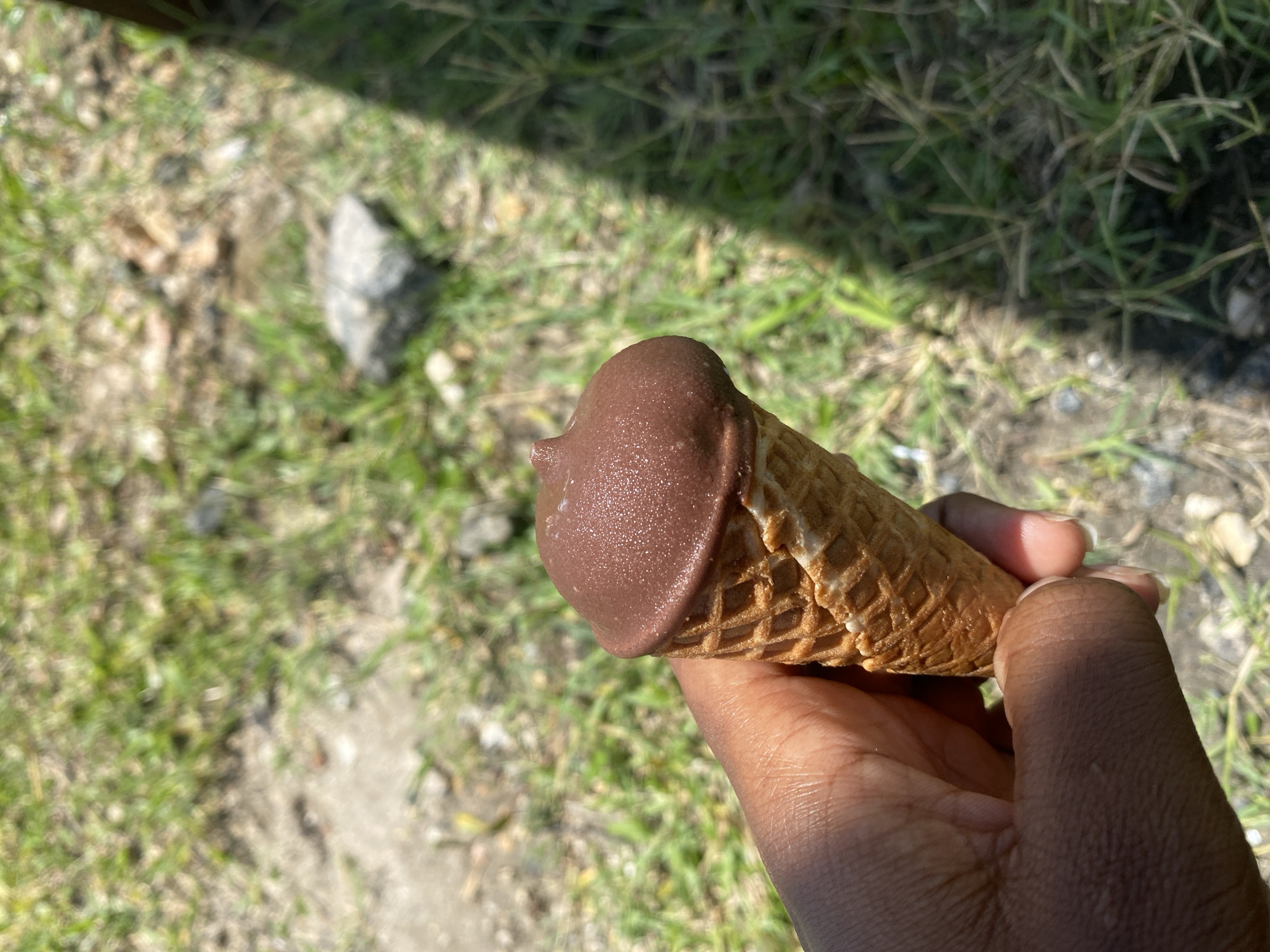 The image presents a close-up, eye-level shot of a person's hand holding a chocolate-dipped ice cream cone. The ice cream features a smooth, medium-brown chocolate shell covering the top, showing a slight sheen and texture from what appears to be tiny sugar crystals or solidification patterns. The chocolate looks somewhat melted, suggesting warm conditions. The cone is a classic waffle cone, with visible grid patterns.
The hand holding the cone has a dark skin tone, with fingers curled around the cone, indicating a firm but relaxed grip. Strong, natural sunlight illuminates the scene, casting bright highlights on the ice cream and the hand, and creating a distinct, elongated shadow that diagonally crosses the upper background. This strong lighting suggests a sunny outdoor setting, likely during midday or early afternoon.
The background is unfocused but reveals an outdoor environment consisting of patches of green and dry grass, interspersed with areas of bare dirt and small stones. The overall impression is a casual outdoor moment. Given the location context of Dar es-Salaam, Tanzania, the bright sunlight and the melting ice cream are consistent with the city's warm, tropical climate. There is no visible text in the image.