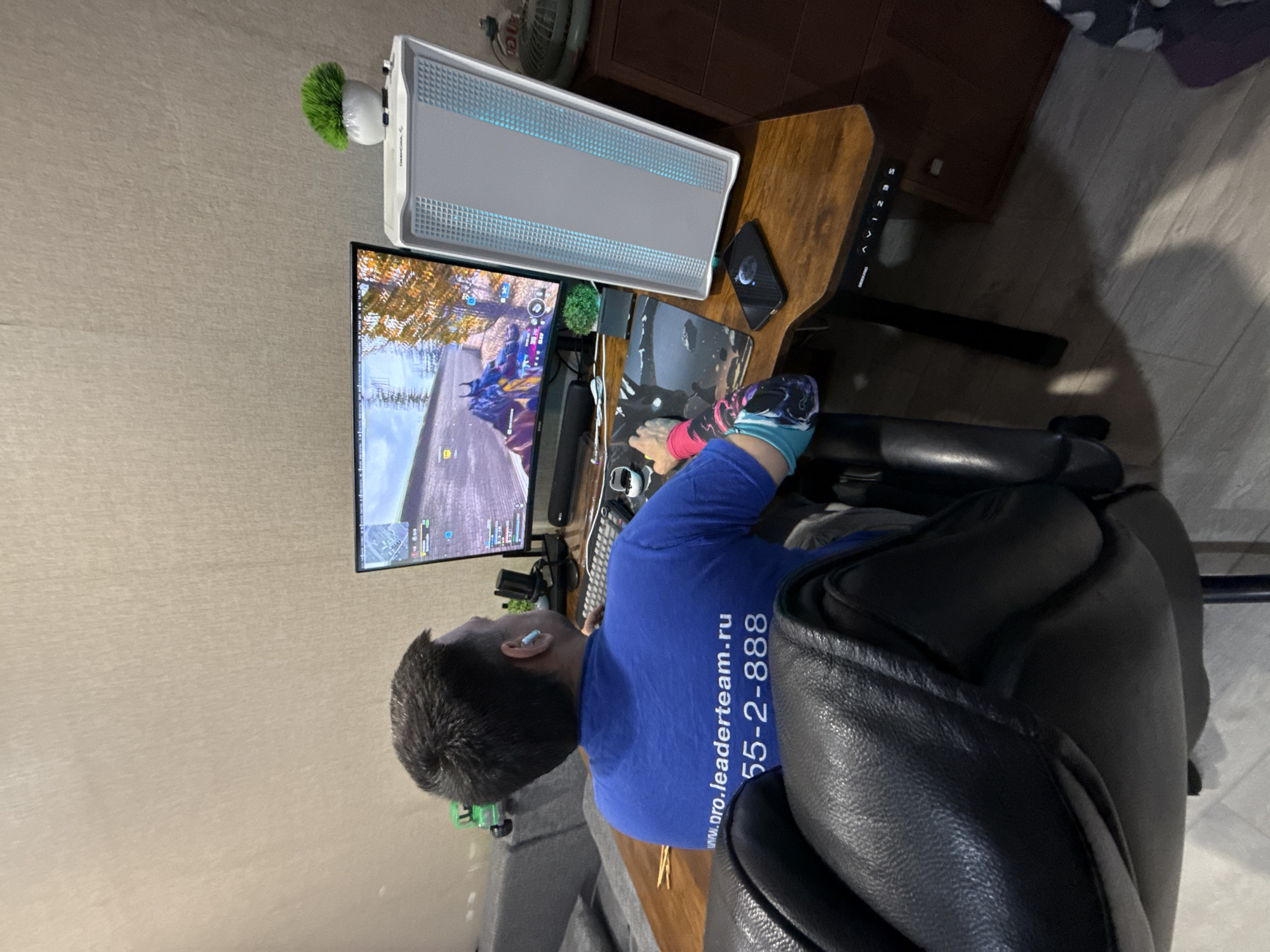 The image shows a person sitting in a black leather chair at a desk, playing a video game on a monitor. The person is wearing a blue t-shirt with text "www.pro.leaderteam.ru 55-2-888". On the desk is a computer tower, a mouse, a keyboard, and a smartphone. A small green plant is positioned near the monitor. A wooden desk is visible with height adjustment buttons. A wooden cabinet is in the background. The location is unspecified, and the city of Rusanovo, Russia is not identifiable within the image.