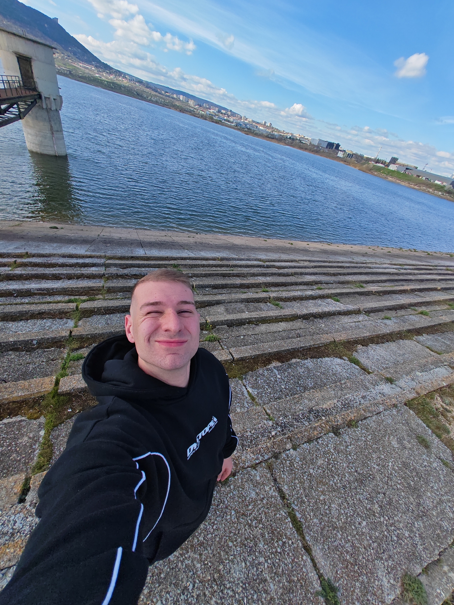 A man is taking a selfie on a stepped concrete embankment next to a body of water. He is wearing a black hoodie with white stripes and the word "DYTOPIA" on it. The man is smiling and winking with one eye. Behind him is a large body of water, and beyond that, a city skyline. To the left of the man and closer to the water is a concrete structure with a metal walkway. The sky is blue with scattered white clouds. The location appears to be in the city of Unknown, Unknown.