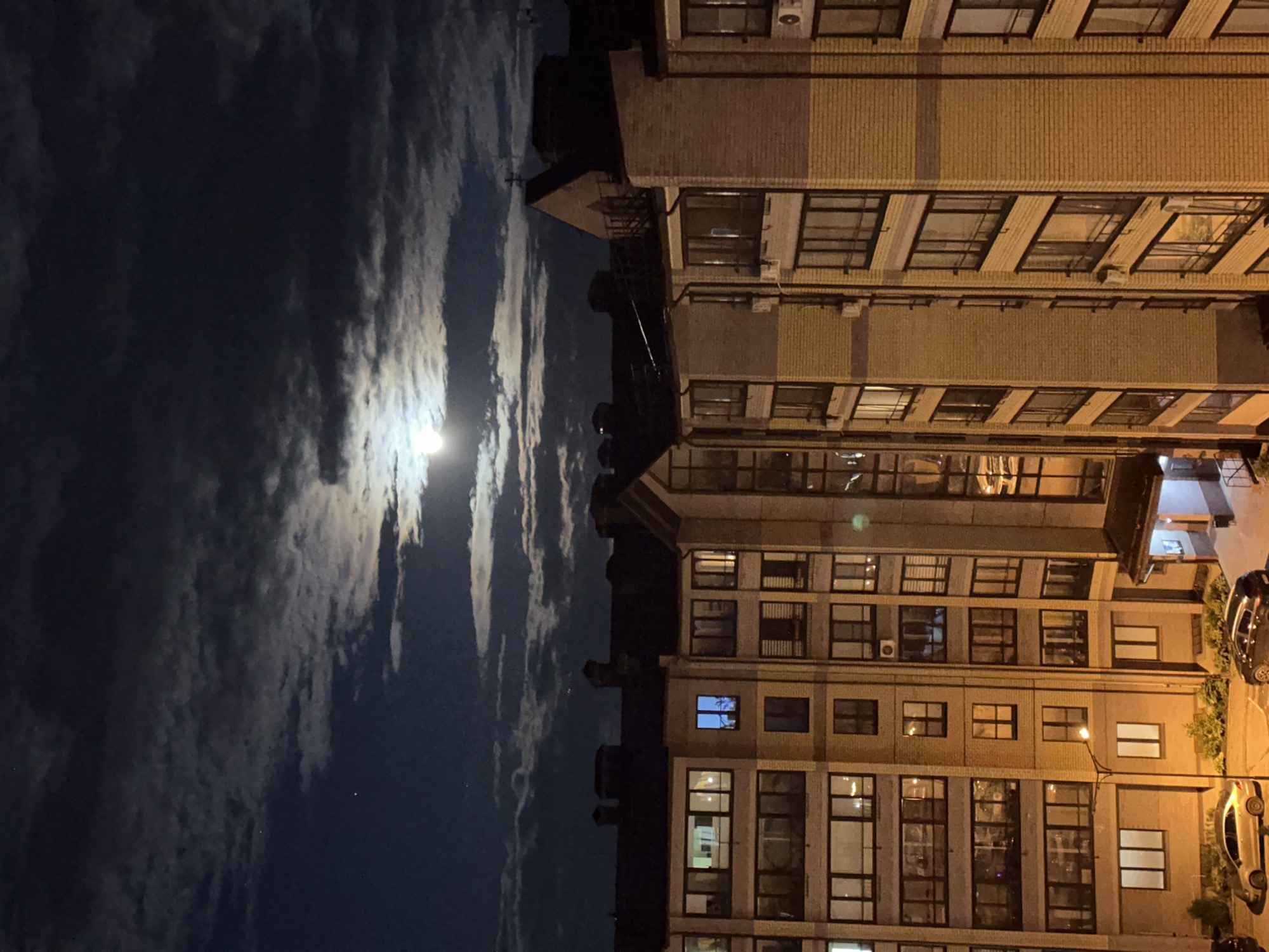 The image shows a building at night in Vladimir, Russia. The sky is dark with clouds and a bright moon. The building is multi-story and lit from the street. Several cars are parked in front of the building.