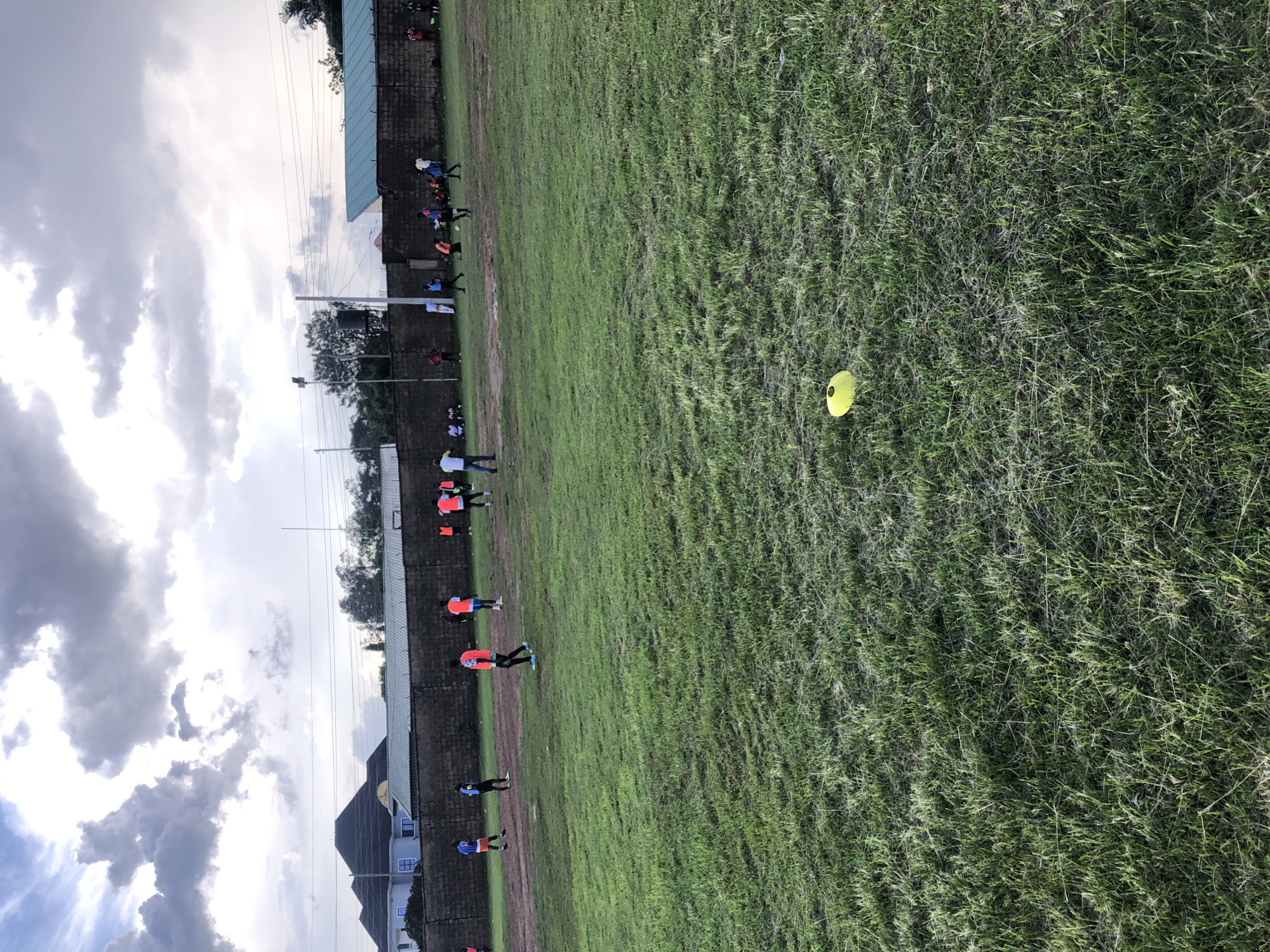 A group of people are gathered on a grassy field, likely a sports field, in what appears to be an outdoor training session. The individuals are dressed in athletic wear, with several wearing bright orange and blue vests, suggesting a team or organized group activity. Some individuals are actively engaged in what looks like drills or exercises, with one person kicking a yellow marker cone in the foreground.
The setting is a large, open field covered in green grass, with a brick wall running along one side. Beyond the wall, there are buildings and trees, and utility poles with wires are visible against a cloudy sky. The time of day appears to be late afternoon or early evening, given the diffused light and dramatic cloud formations. The weather seems overcast, with a mix of dark and bright clouds.
The image is oriented vertically, with the field stretching from the bottom of the frame upwards towards the sky. The overall mood is one of focused activity and potentially competitive sport. There is no visible text in the image. The location context of Jos, Nigeria, provides a geographical setting for this scene of an outdoor sporting event.