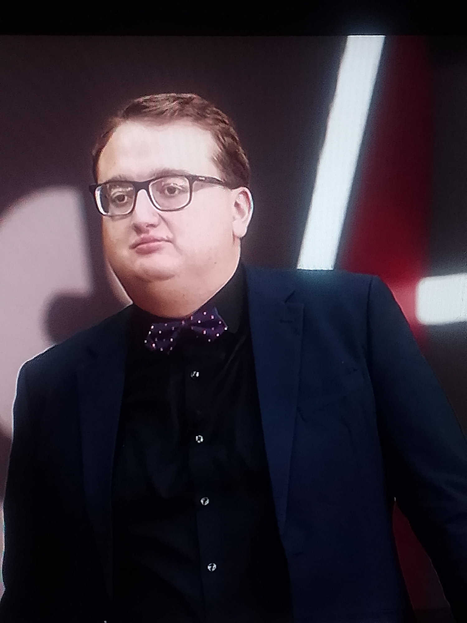 A man with brown hair wearing glasses, a dark shirt, and a bow tie stands in front of a blurred background. He is also wearing a dark blazer. The city and location are unknown.