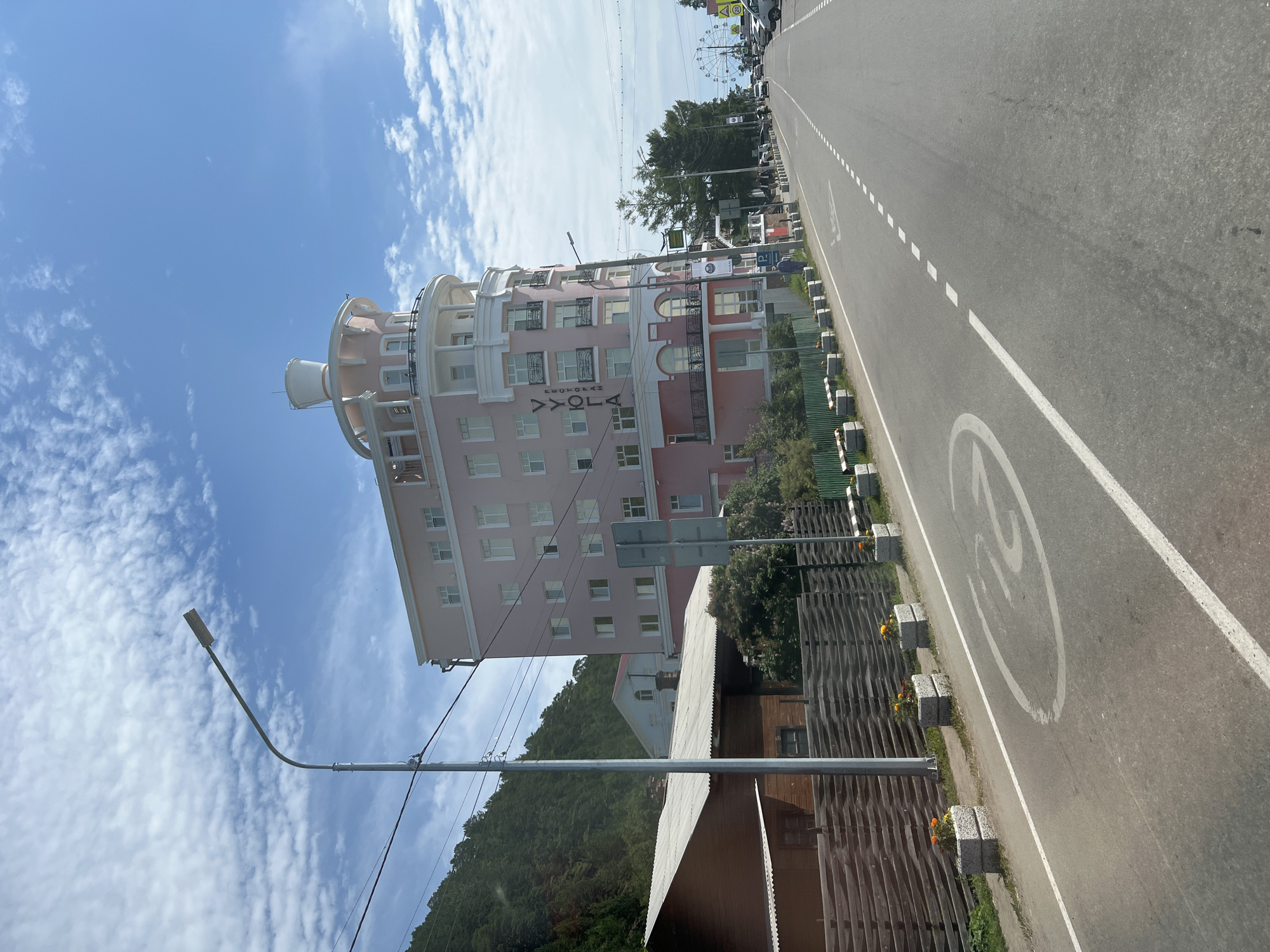 This image depicts a street scene in Listvyanka, Russia. A distinctive pink, multi-story building with a rounded facade and a white turret-like structure on top is prominent. The building has many windows and balconies. Text reading "УЮГЛА" and "РЕСТОРАН" is visible on the building's facade. To the left of the building, a dark wooden structure with a corrugated metal roof is situated behind a wooden fence. A street lamp stands in the foreground, near a paved road. The road has white lane markings and a circular symbol painted on it, possibly indicating a traffic regulation. Flower planters with orange flowers are placed along the roadside. In the background, a forested hill is visible to the left, and further down the road, there are other buildings, trees, parked cars, and a Ferris wheel. The sky is blue with scattered white clouds. The image appears to be taken from a moving vehicle.