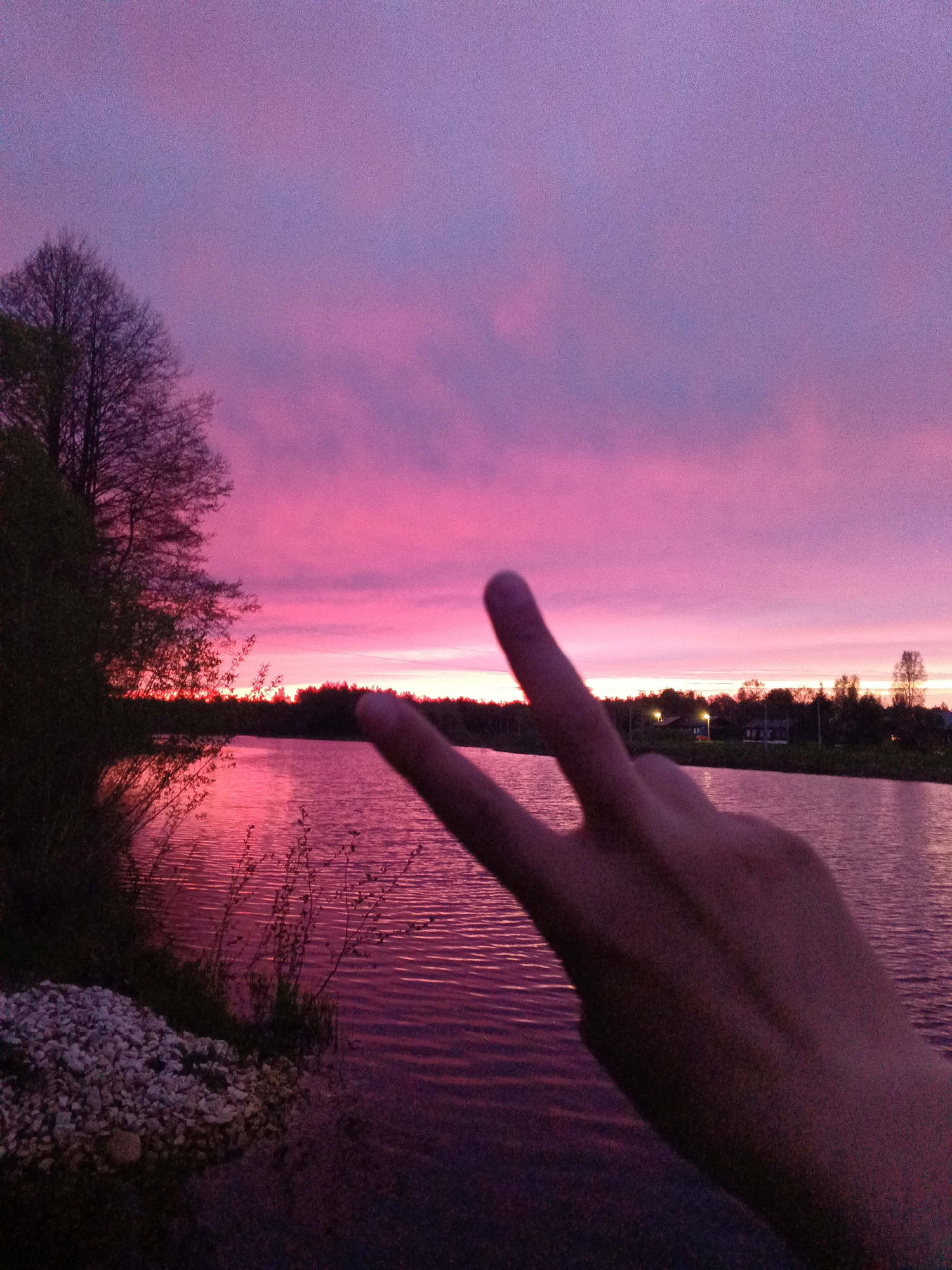 A scenic shot captures a pink and purple sunset over a body of water, presumably a lake or river, in Красный Маяк, Russia. Trees and other vegetation are visible along the shoreline. In the foreground, a hand is making a peace sign, partially obscuring the view. The scene suggests a tranquil or picturesque setting.