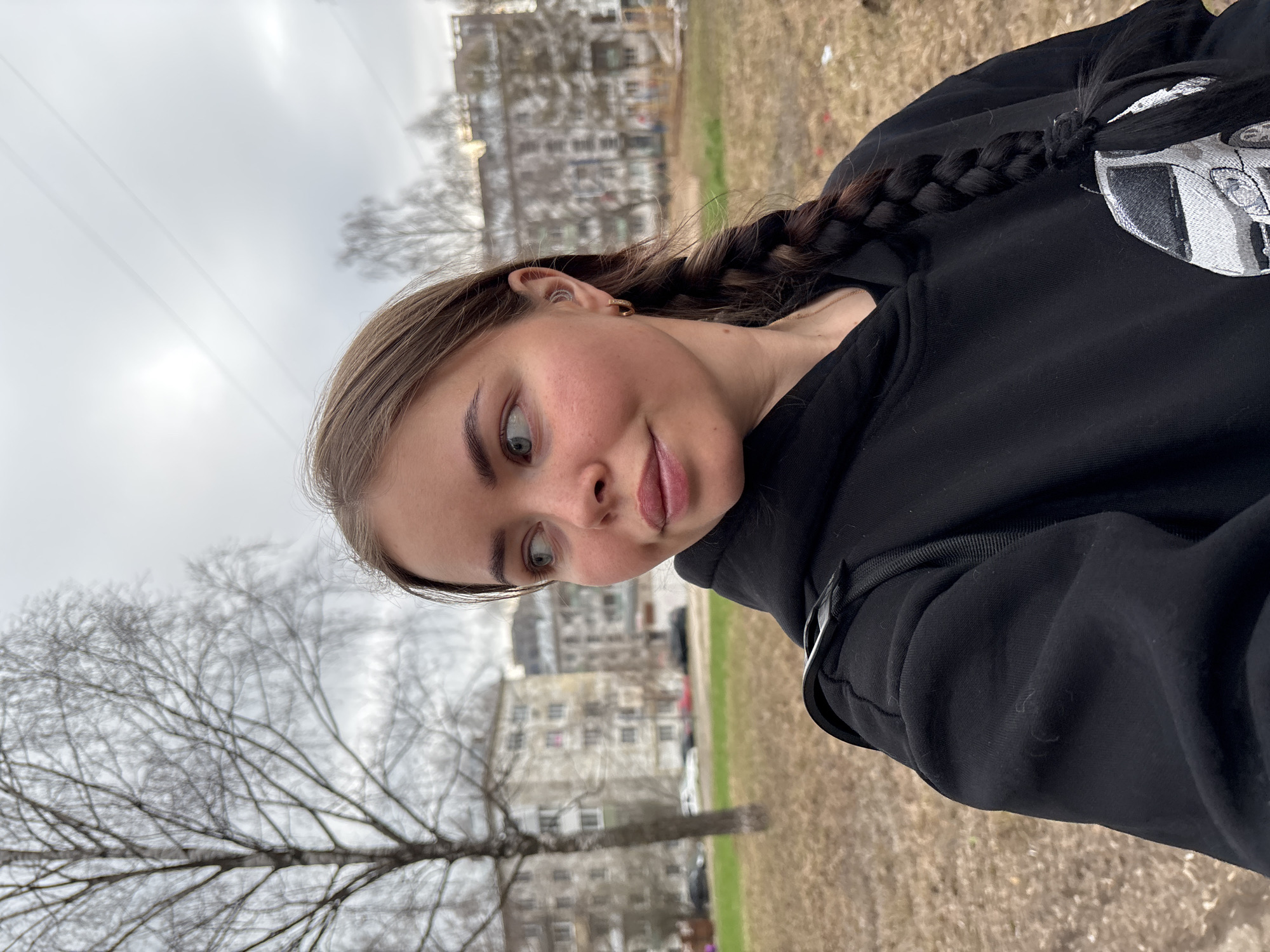 A young woman is pictured outdoors in what appears to be a park or green space. The woman has light brown hair with a dark braid, light-colored eyes, and fair skin. She wears a black hooded sweatshirt with a design on the shoulder. The background features leafless trees, dry grass, and several multi-story buildings, characteristic of urban residential architecture. The location is potentially Izhevsk, Russia.