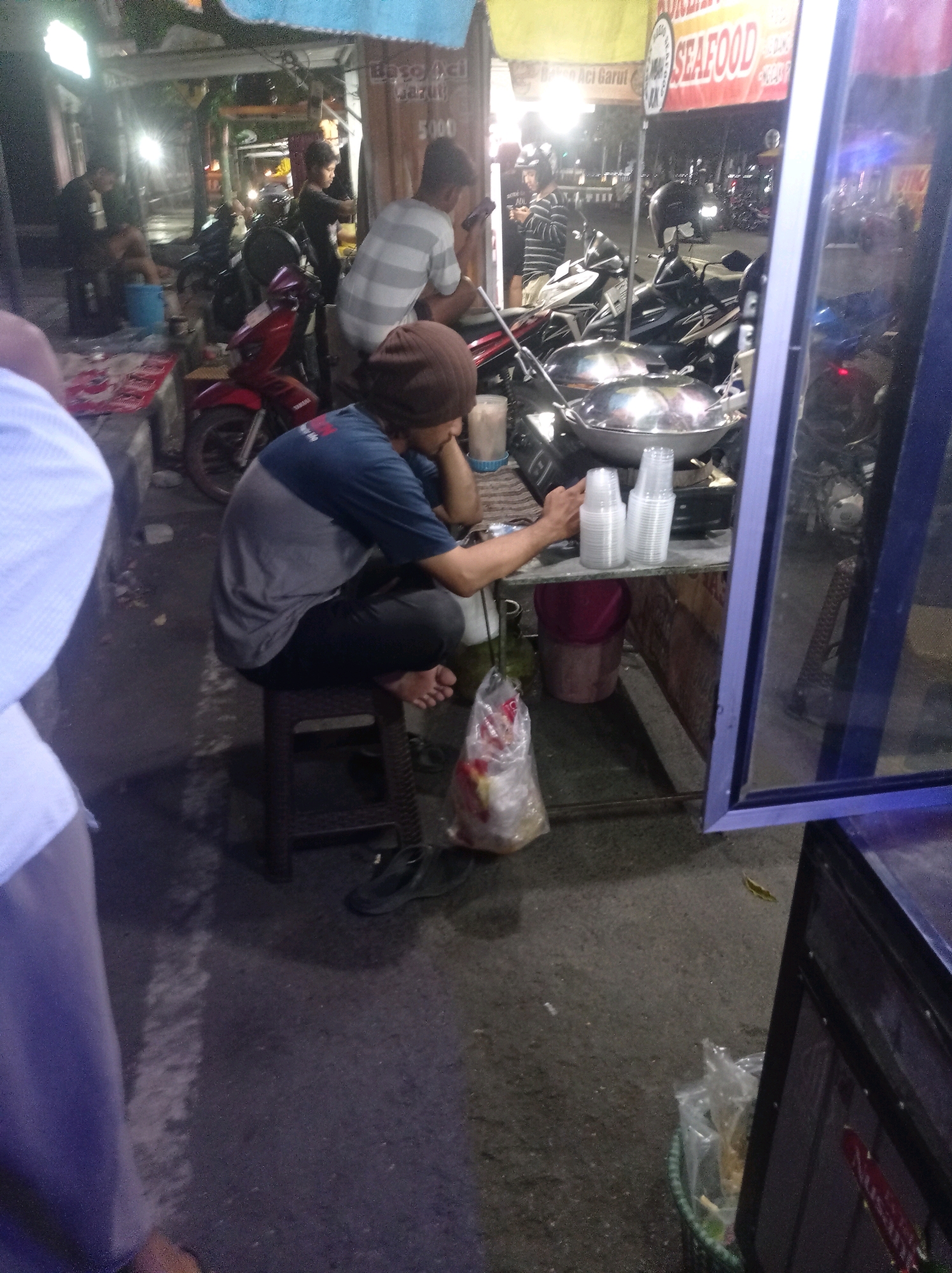 The image shows a street scene at night in Tuban, Indonesia, likely a food stall area. A man wearing a dark brown beanie and a grey and blue t-shirt is sitting on a plastic stool, facing away from the camera and looking down at his phone or a device. He is barefoot. In front of him, on a metal counter, are two woks, plastic cups, and other items associated with food preparation or serving. To the left of the counter, a plastic bag filled with food items hangs from a hook. The background shows a street with several motorcycles parked, a food stall with a red and white sign that reads "SEAFOOD" in large letters, and a blue and purple framed display case. Lights from the stalls and street illuminate the scene. The ground appears to be asphalt.