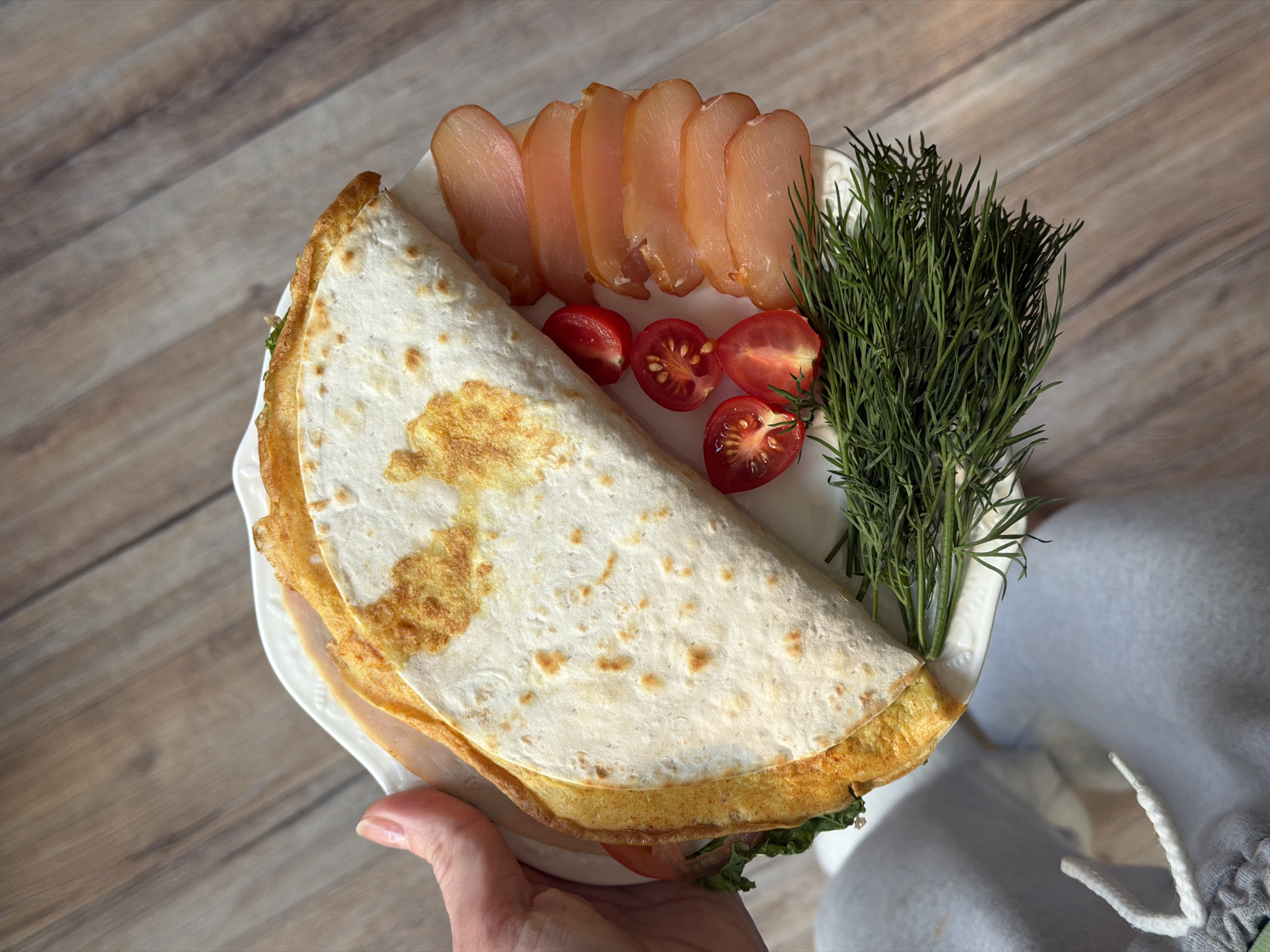 A person holds a white plate of food over a wooden surface. The plate contains a folded quesadilla with browned edges, several slices of light-colored meat, halved cherry tomatoes, and a bunch of dill. The plate is positioned slightly above the person's hand and leg, with the leg appearing to be clad in light gray sweatpants. The background consists of a wooden surface that resembles a table or floor. The location is Perm, Russia.