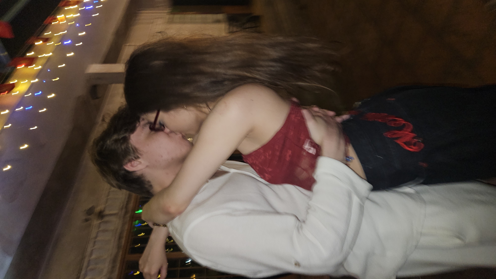 A blurred image shows a couple kissing. A man wearing a white long-sleeved shirt is lying down. A woman with long brown hair, wearing a red top and black pants, is on top of him. They are kissing. The location appears to be outdoors. Lights are visible in the background. The city of Nha Trang, Vietnam, is assumed but unconfirmed by visual evidence.