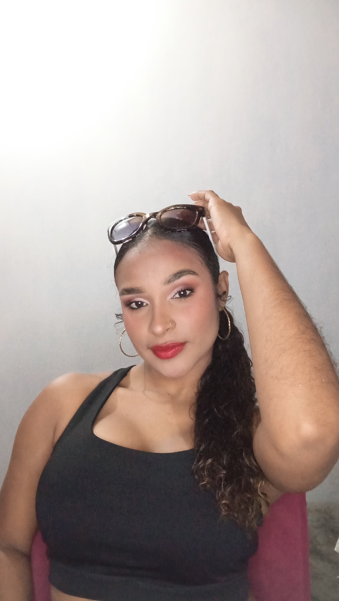A woman is posing for a selfie. She is wearing a black tank top, gold hoop earrings, and has sunglasses on her head. Her hair is pulled back in a ponytail. She has red lipstick on and a nose ring. The background is a plain white wall. The location is unknown.