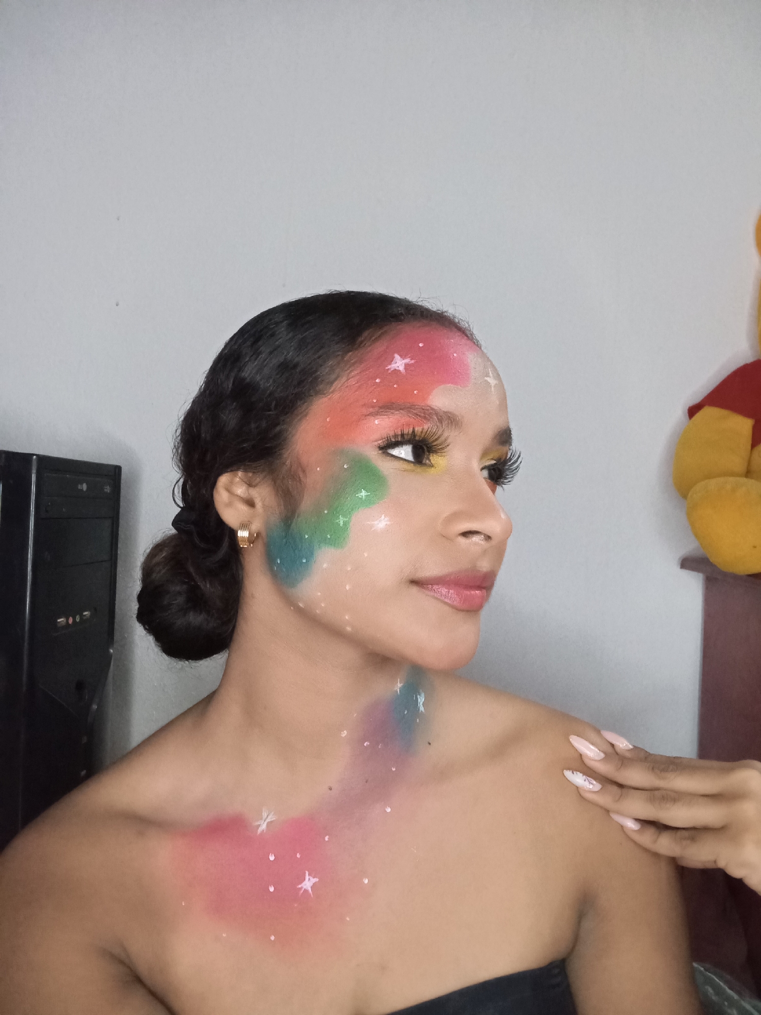 A young woman with dark hair pulled back into a bun and a fair complexion is pictured from the chest up, facing right with her head turned slightly towards the camera. She has colorful, abstract makeup resembling a galaxy painted on her face and chest, including shades of pink, green, and blue, with small white star shapes scattered across it. She wears gold hoop earrings and false eyelashes. Her lips are lightly tinted pink. Her right hand is visible, gently touching her right shoulder. She has long, manicured nails with a light pink polish and a simple white design.
In the background, a grey wall is visible, along with the side of a black computer tower and a stuffed Winnie the Pooh toy on a wooden shelf or surface. It is not possible to determine if the location is the city of Juan Griego, Venezuela, based on the image.