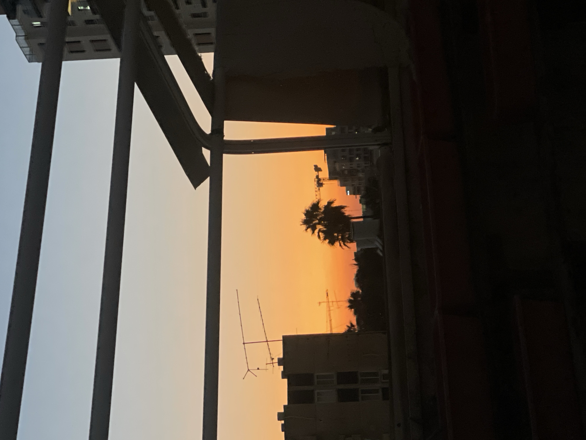 An outdoor, possibly rooftop, view of a sunset in Petah Tikva, Israel. The sky transitions from a pale blue at the top to a deep orange towards the horizon. Silhouetted against the sky are several buildings, including a multi-story building with visible windows on the left, and another taller building on the right. A palm tree is visible in the middle distance, and construction cranes can be seen beyond the buildings. In the foreground, dark, tiered seating is visible, suggesting the location might be a stadium or amphitheater. The view is partially obstructed by dark, horizontal beams and a metal structure that forms a frame around the scene.