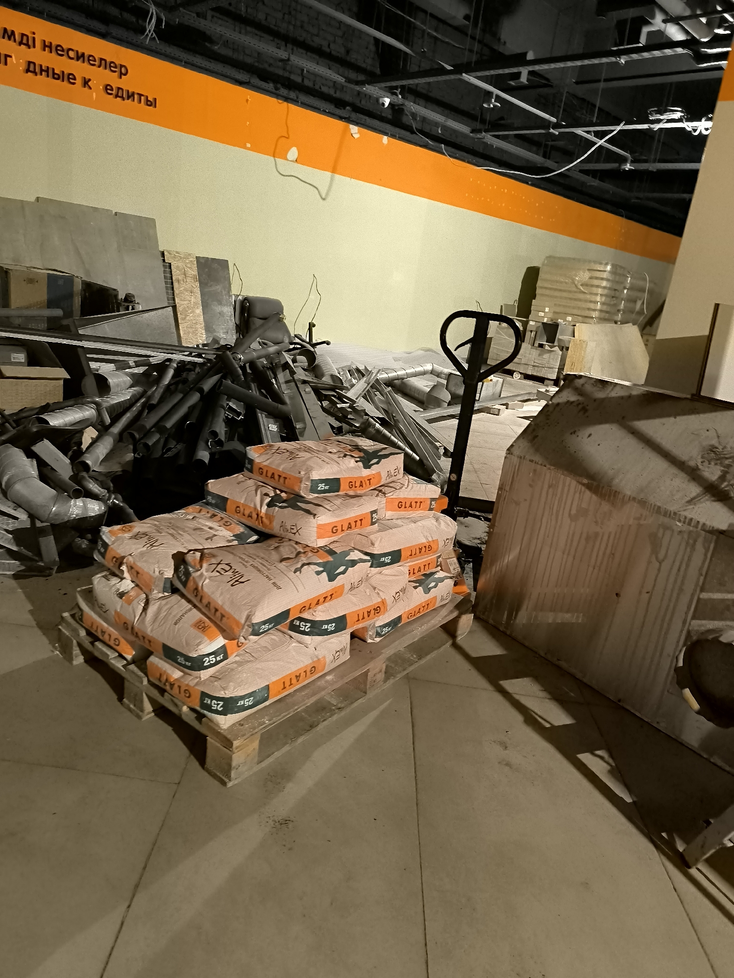 The image depicts a dimly lit interior space that appears to be under construction or renovation. In the foreground, several stacks of bagged material, likely construction supplies such as cement or dry mix, are arranged on a wooden pallet. The bags are predominantly light brown with orange and green branding that reads "GLATT" and "ALEX". To the left of the bags, a pile of metal pipes and ductwork is visible, suggesting construction or plumbing work. A black manual pallet jack stands near the stacks of bags. In the background, various construction materials and debris are scattered, including large sheets of what appear to be drywall or insulation. The walls are painted a pale yellow or green, and an orange stripe runs along the top portion of the wall. The ceiling is a dark, industrial structure with exposed pipes and wiring. The floor is a concrete surface with visible seams. The overall impression is one of an unfinished or storage area within a building. There are no people visible in the image. This scene does not provide any specific indicators of its location within the city administration of Qarağandı city, Kazakhstan.