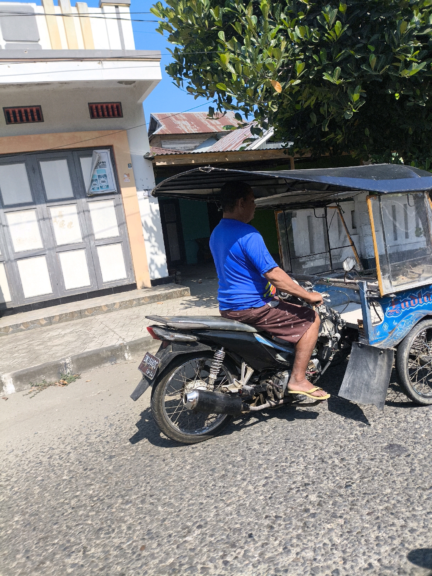 The image captures a man operating a three-wheeled motorized vehicle, resembling a tuk-tuk or a motorcycle-drawn carriage, on a street in Pentadu Barat, Indonesia. The man, wearing a blue t-shirt and brown shorts, is seated on the motorcycle portion, facing right and holding the handlebars. The vehicle has a black canopy and a clear plastic enclosure at the front, with visible orange trim. The background shows a building with a light-colored facade and dark gray double doors, as well as a rooftop with corrugated metal. Green foliage from a tree overhangs the scene. The street is paved with asphalt, and a concrete sidewalk runs alongside the building. The license plate of the vehicle is partially visible and appears to have alphanumeric characters. The lighting suggests it is daytime, with a clear blue sky.