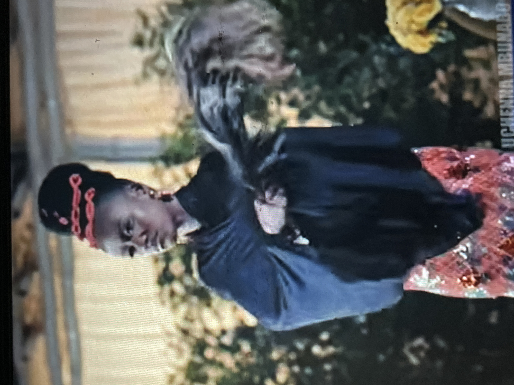 The media file displays an image, rotated 90 degrees clockwise, depicting a person, likely a woman, in what appears to be a performance or dance. The image quality is low, showing blurriness, pixelation, and artifacts consistent with a photograph of a screen.
The central figure, with dark skin, is adorned in a dark, possibly navy or black, draped top or shawl. Her lower attire is a vibrant, patterned skirt featuring colors like red, pink, orange, green, and white. Her hair is elaborately styled, with distinct red, possibly beaded or braided, adornments encircling her head. Long, dark hair strands are captured in mid-motion, flowing upwards and to the left, indicating active movement. Her facial expression is one of focus or concentration.
The background is largely blurred, showing green foliage, a light-colored, possibly fabric or wooden, structure to the left, and a blurry yellow object, possibly floral decor, in the upper right. A very indistinct, blurry figure or object is also visible behind the main subject's head.
In the upper right portion of the image, oriented vertically, the name "UCHENNA MBUKADO" is clearly legible. The overall setting suggests a daytime event, potentially outdoors or in a well-lit celebratory venue, aligning with the cultural context of Abuja, Nigeria. The attire and dynamic pose strongly imply a cultural or celebratory performance.