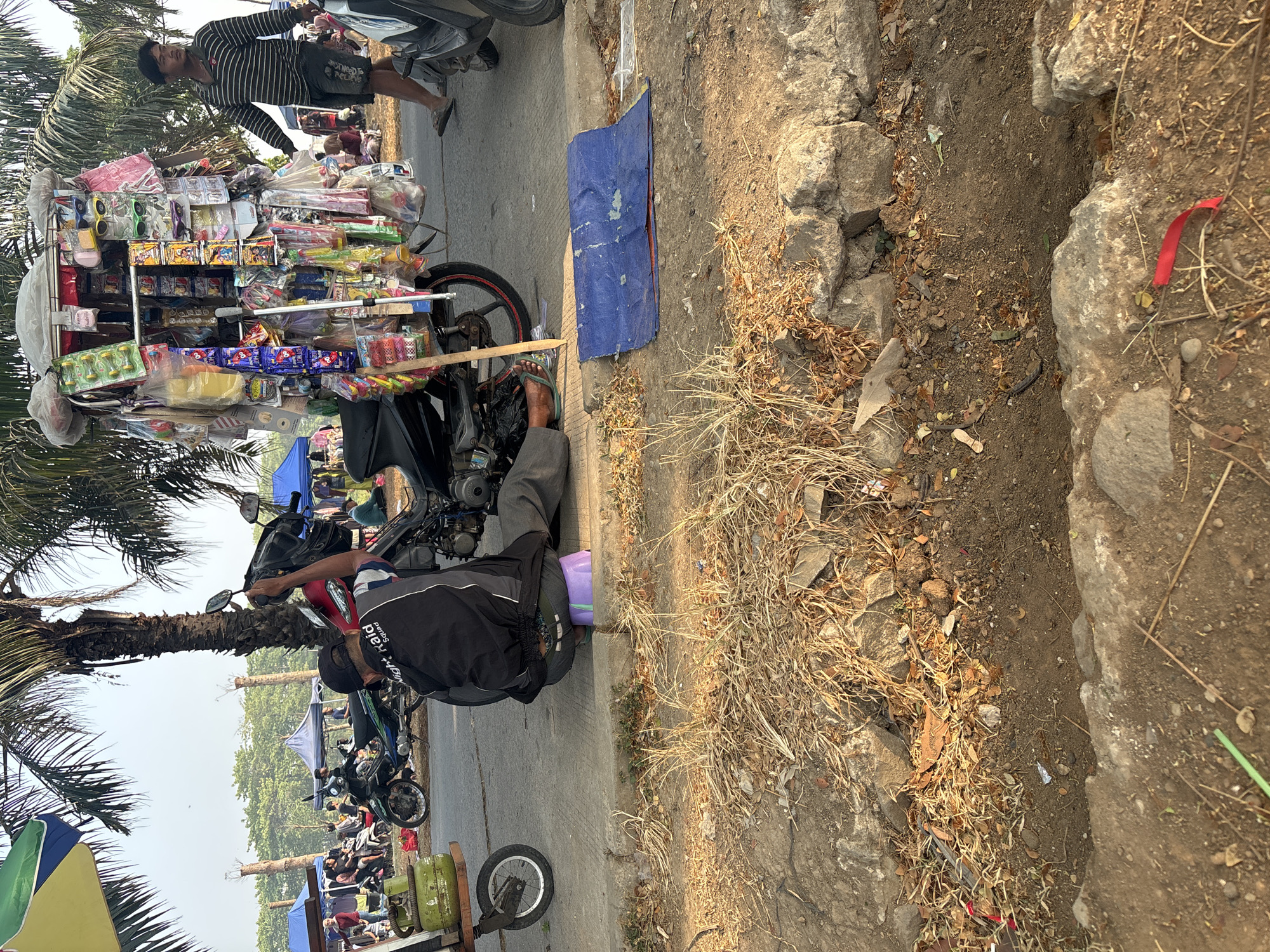 A man is sitting on a purple stool on a sidewalk next to a motorcycle laden with merchandise. He is wearing a black jacket with "Night Raid Squad" printed on the back and grey pants. The motorcycle is parked next to a sidewalk, with its front wheel on the sidewalk. A cart with a green gas tank is to the left of the motorcycle. In the background, there are several blue tents and other motorcycles. A man wearing a striped shirt and shorts is walking away from the motorcycle. The ground is dirt and dry grass, with some rocks visible. The scene appears to be outdoors in a market or roadside setting. The location is Telagamurni, Indonesia.