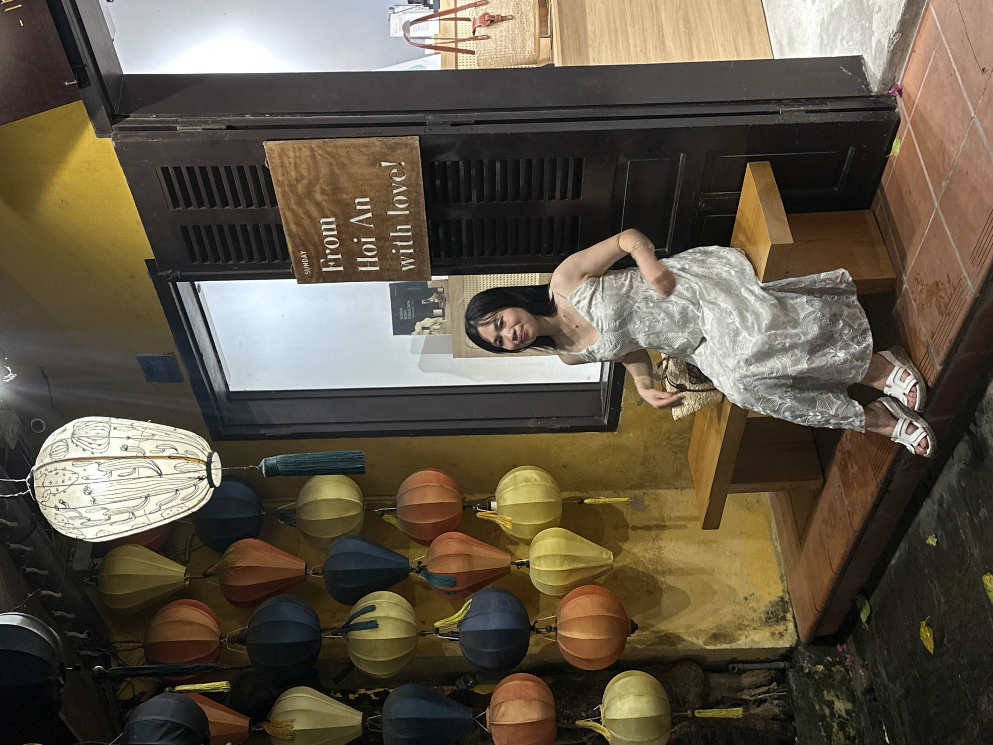 A young Asian woman is seated on a wooden bench in front of a dark wooden door with louvers. She is wearing a light-colored, lace-patterned dress and white sandals. The woman is smiling and looking towards the camera. Behind her, a sign reads "From Hoi An with love!". The scene appears to be outdoors, possibly at dusk or nighttime, given the lighting. A large, white lantern with a detailed drawing of cacti and other desert plants hangs on the left side of the frame. Below it, a string of colorful, round lanterns, in shades of orange, yellow, and blue, are displayed. The wall behind the lanterns is a textured yellow. The ground is dark and appears wet in places. The overall atmosphere suggests a vibrant, possibly tourist-oriented location in Hoi An, Vietnam, characterized by its traditional lanterns and charming ambiance.
