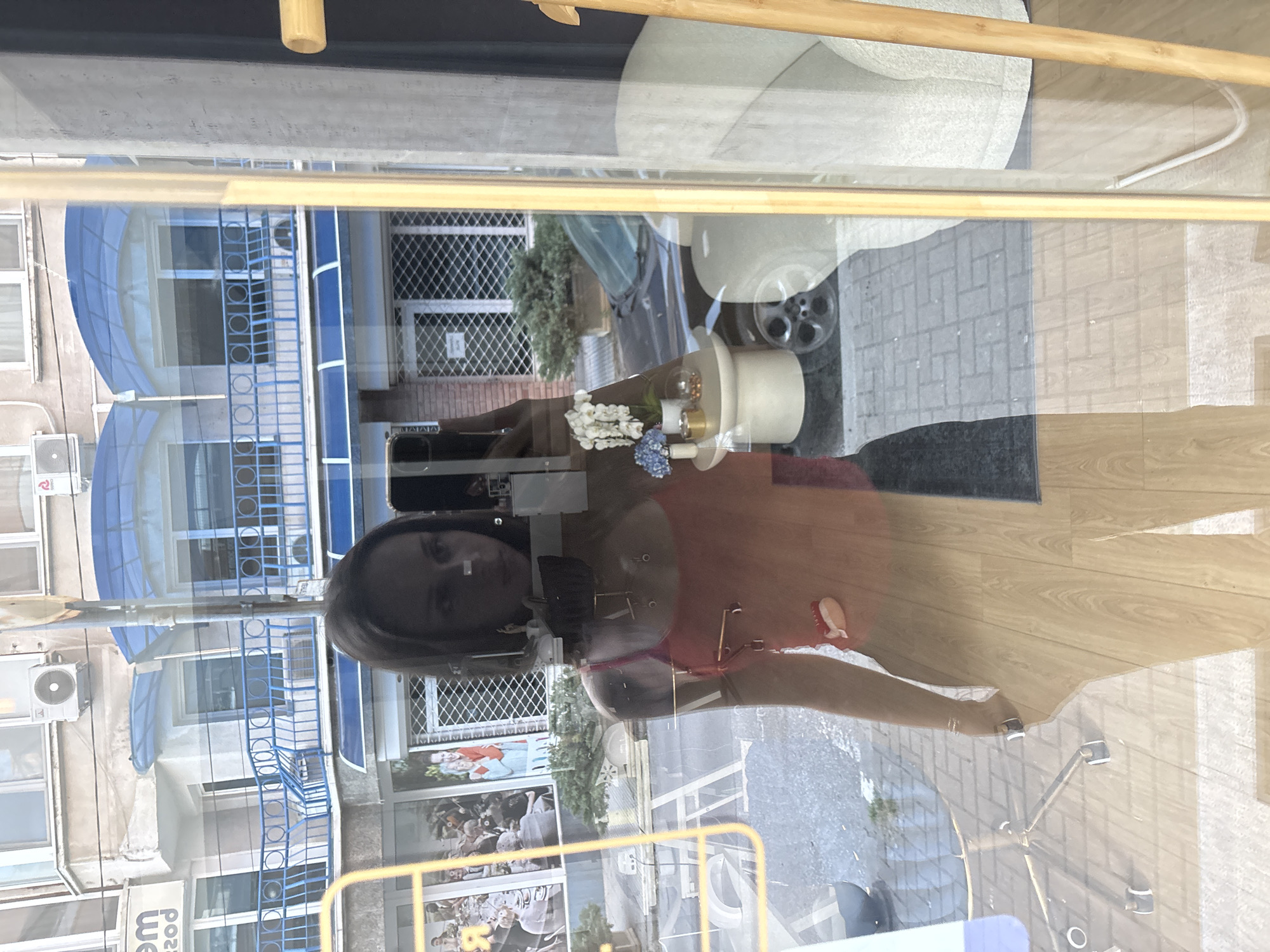 A woman's reflection is seen in a shop window. The reflection captures a street scene in Sofia, Bulgaria, featuring a building with blue awnings and metal railings, air conditioning units, and posters or photographs displayed on a wall. Inside the shop, a display includes a white lamp, flowers, and a chair. The flooring is light-colored wood-effect laminate.