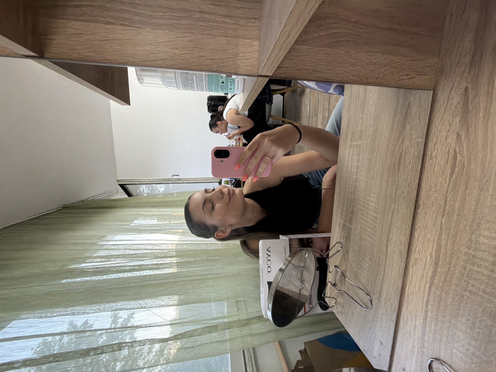 A mirror selfie of a young woman is reflected onto a vanity tabletop. The woman is holding a pink-cased mobile phone with her right hand and has fair skin and brown hair pulled back from her face. She is wearing a black sleeveless top. On the vanity table next to her is a makeup mirror on a stand and a cosmetic product box. The background behind the vanity shows a room with green curtains and a window with a partial view of the outside. A second woman is visible in the mirror, sitting and applying makeup. The location could be Sofia, Bulgaria but further details are needed to confirm.