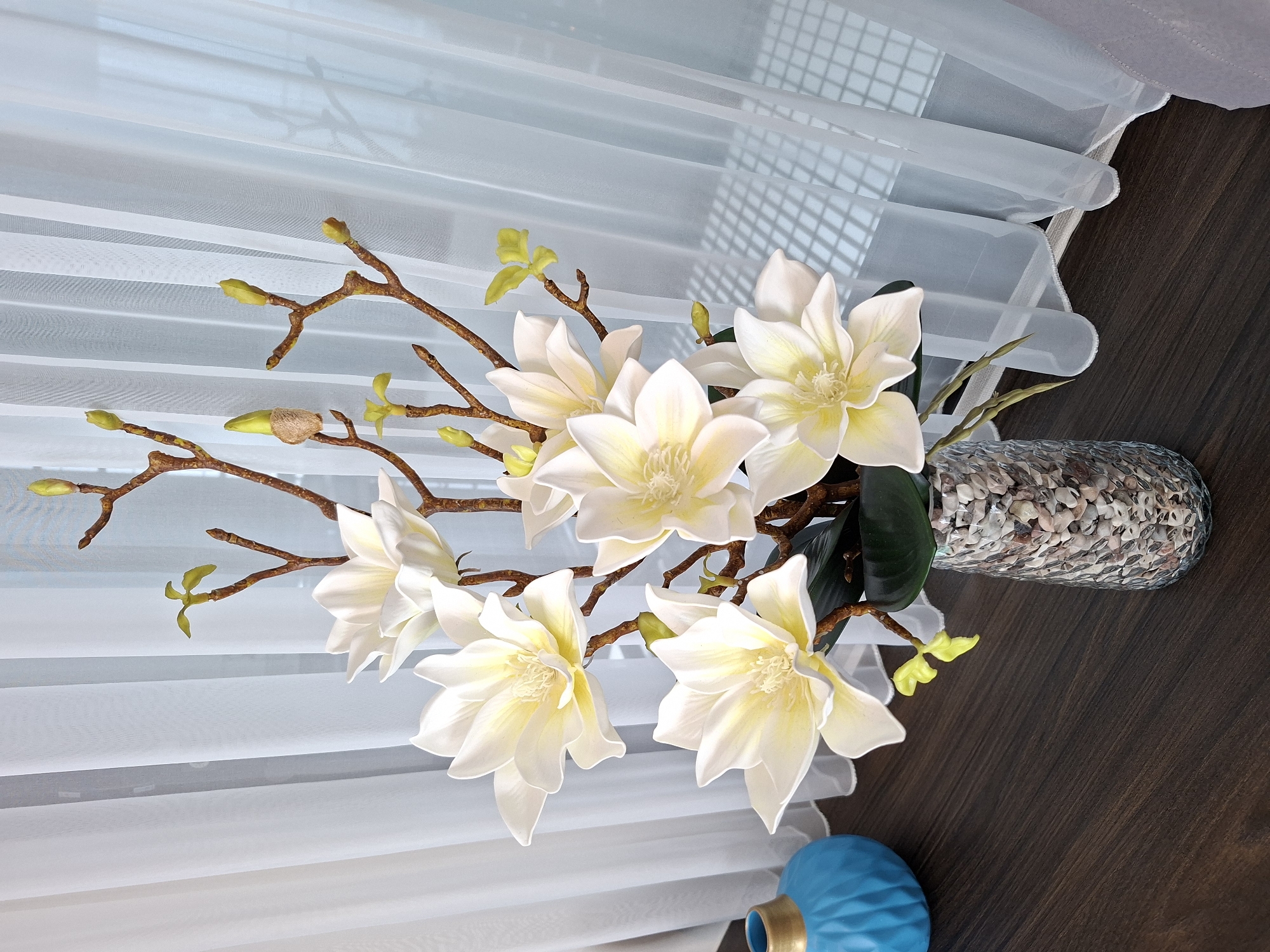 A still life photograph showcases a bouquet of artificial white magnolia flowers with yellow centers, arranged in a textured glass vase filled with small pebbles. The bouquet is placed on a dark wooden surface in front of sheer white curtains. To the left of the vase, a portion of a textured, turquoise-colored vase with a golden rim is visible. The background consists of the white curtains and a glimpse of a window grid pattern. There are no people or animals present in the image. The scene appears to be an indoor setting, likely a home or a decorated commercial space. The image does not contain any information to ascertain its location within the city of West Jakarta, Indonesia, or any other specific city.