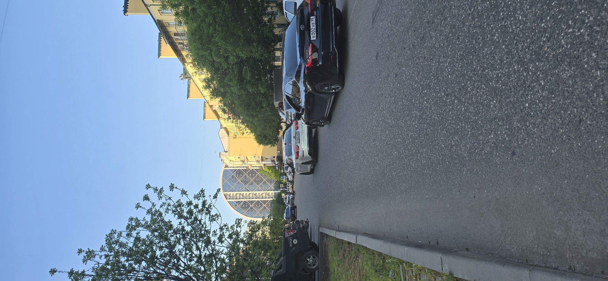 The image shows a street scene in Grozny, Russia, identified by the distinctive Circular House building in the background. The street is lined with parked cars. A row of trees separates the buildings from the road. The sky is clear blue.