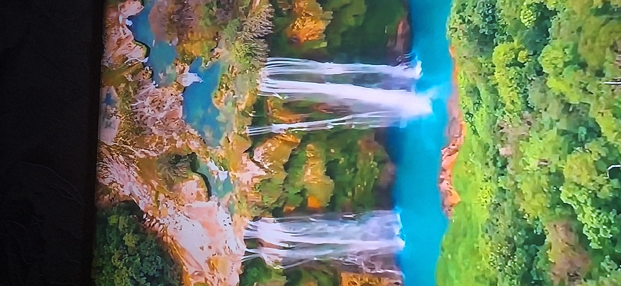 The image depicts a picturesque natural landscape featuring waterfalls cascading into a vibrant turquoise pool. The surrounding environment is lush and green, with dense foliage blanketing the foreground and climbing up the rocky cliffs. The waterfalls themselves appear as thin streams of white water against a backdrop of moss-covered rocks and earthy tones. The sky is a soft blue, suggesting a clear day. The scene conveys a sense of tranquility and natural beauty. This image does not depict any location in the city of Bila Tserkva, Ukraine.