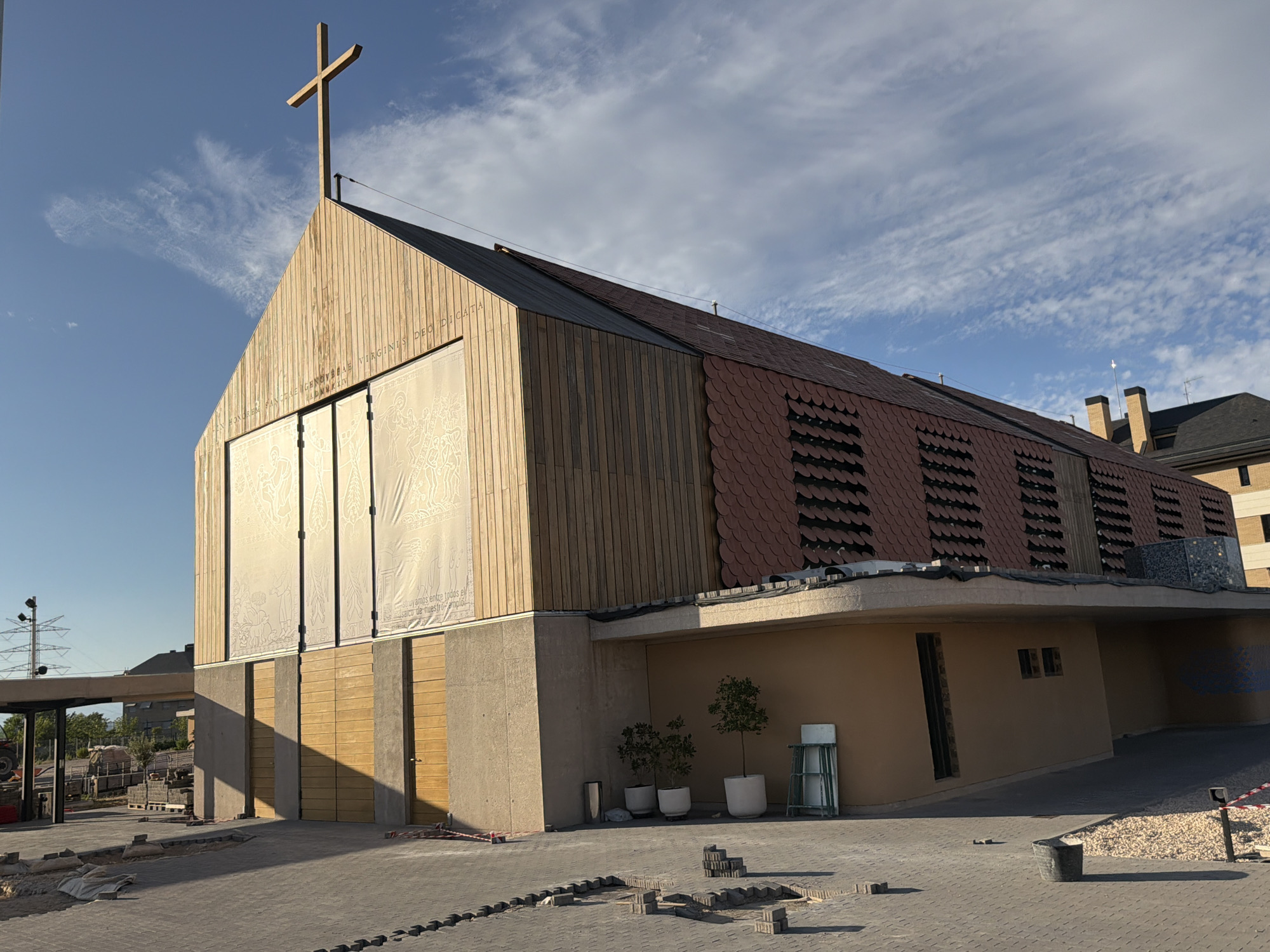 The image shows a modern church building with a prominent wooden facade topped with a cross. The roof is covered with dark red, scaled tiles. The building features large, pale panels with etched designs on the front, below the wooden facade, and several wooden doors on the lower level. The surrounding area appears to be under construction, with paving stones laid out and some materials stacked. Two small trees in white pots and a metal shelving unit are visible near the entrance. In the background, a utility tower and other buildings can be seen under a partly cloudy blue sky. The scene is illuminated by sunlight, casting shadows on the ground. The church is located in Majadahonda, Spain.