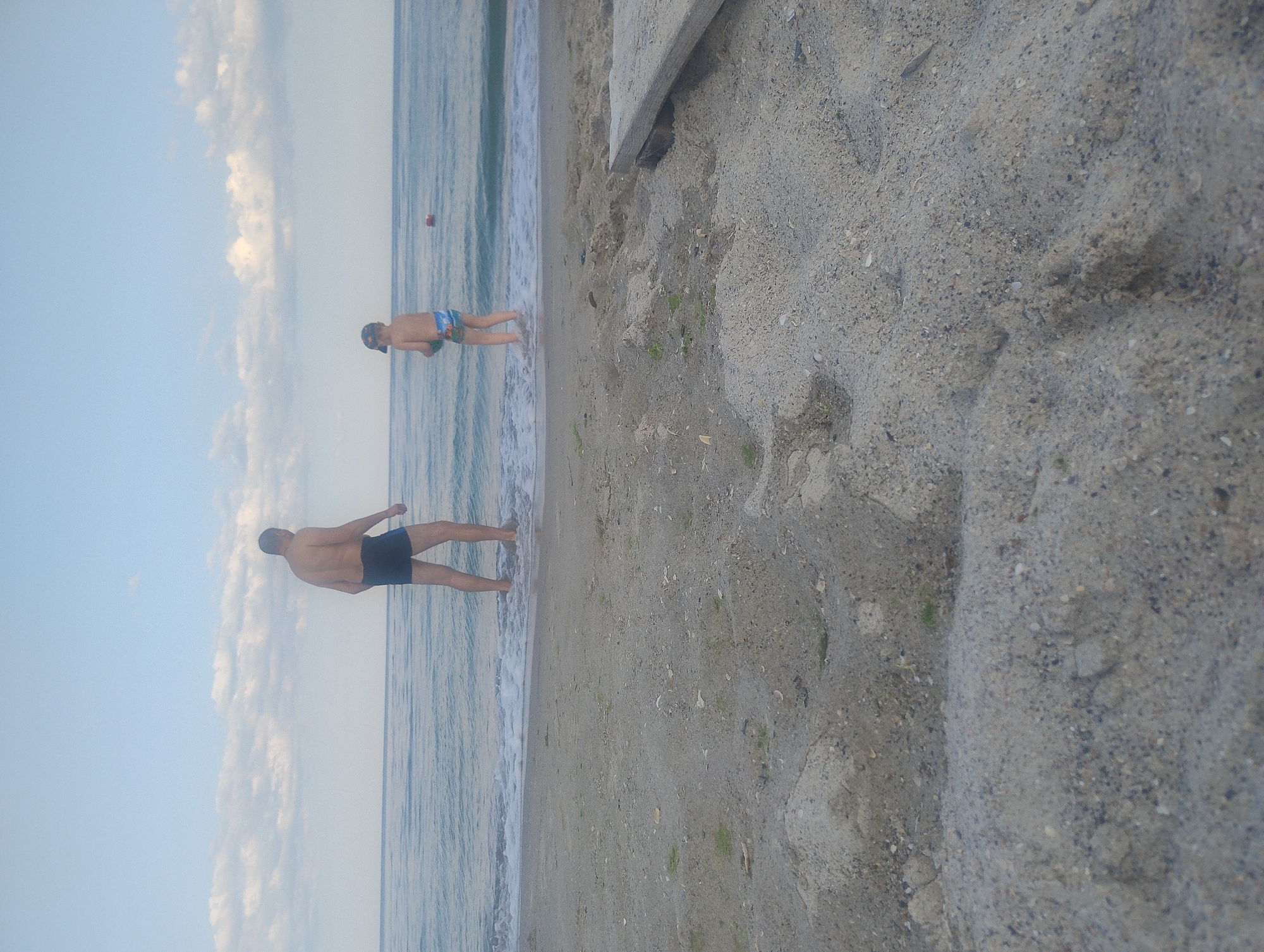 An adult male and a child are standing in shallow water at the edge of a sandy beach. The male is wearing dark swim trunks and is facing away from the camera. The child is wearing swim trunks with a pattern and a baseball cap, and is also facing away from the camera. The water is calm and the sky is partly cloudy. There is a small red buoy in the water in the distance. The sand in the foreground is rough and appears to have shells and debris mixed in. A wooden plank is visible on the right side of the image, partially buried in the sand.